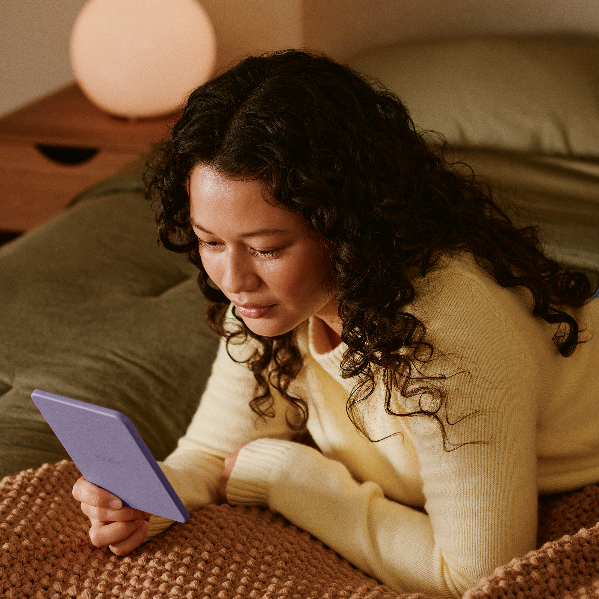 Eine Frau mit dunklen, lockigen Haaren liegt auf einem Bett und liest auf einem lila E-Reader.