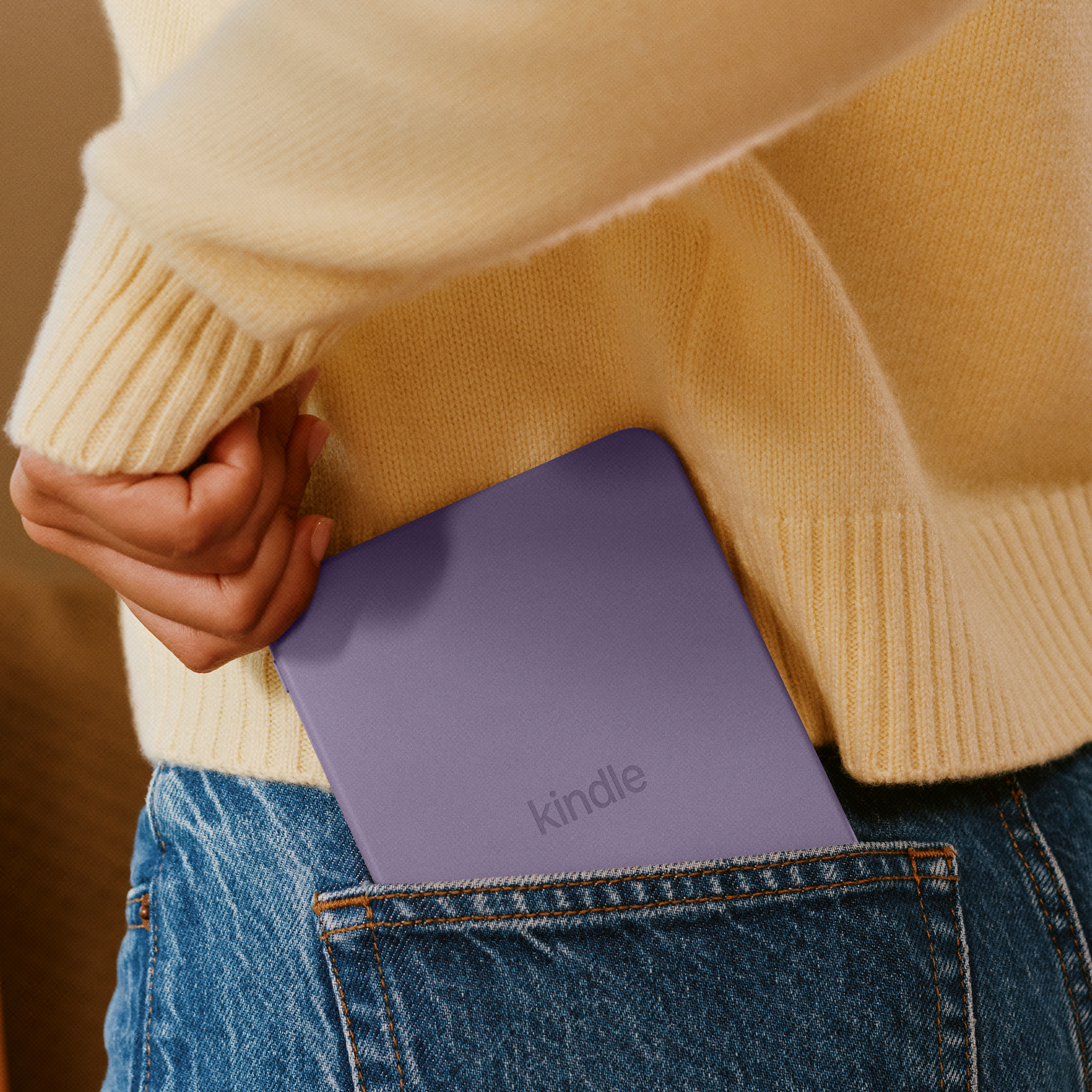 Person in gelbem Pullover und Jeans steckt einen lilafarbenen Kindle in die Gesäßtasche.