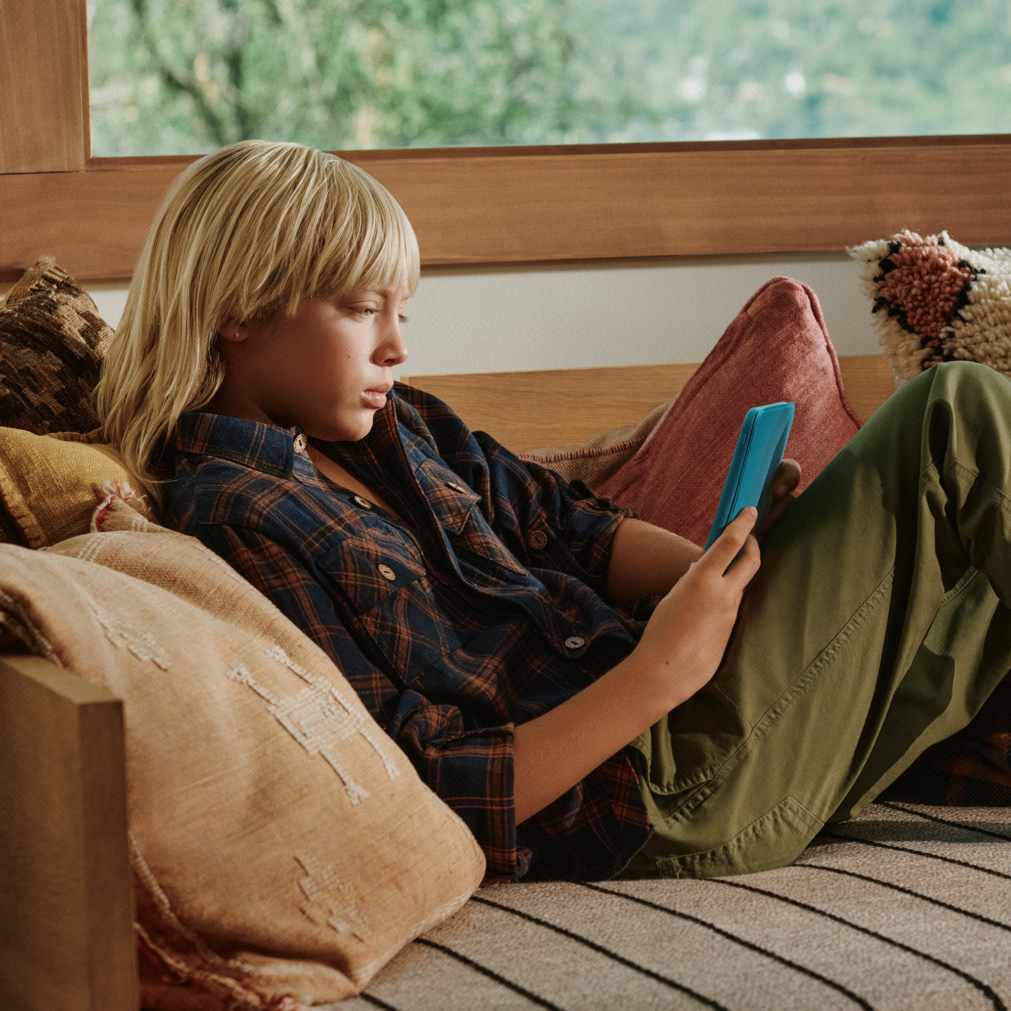Ein blondes Kind mit kariertem Hemd sitzt auf einer Couch und nutzt ein blaues Tablet.