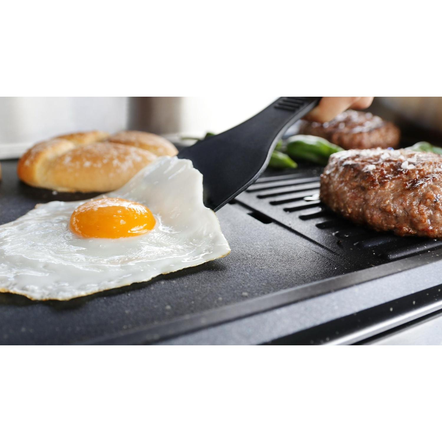 Un uovo al fritto viene girato con una spatola su una piastra calda accanto a hamburger e pane.
