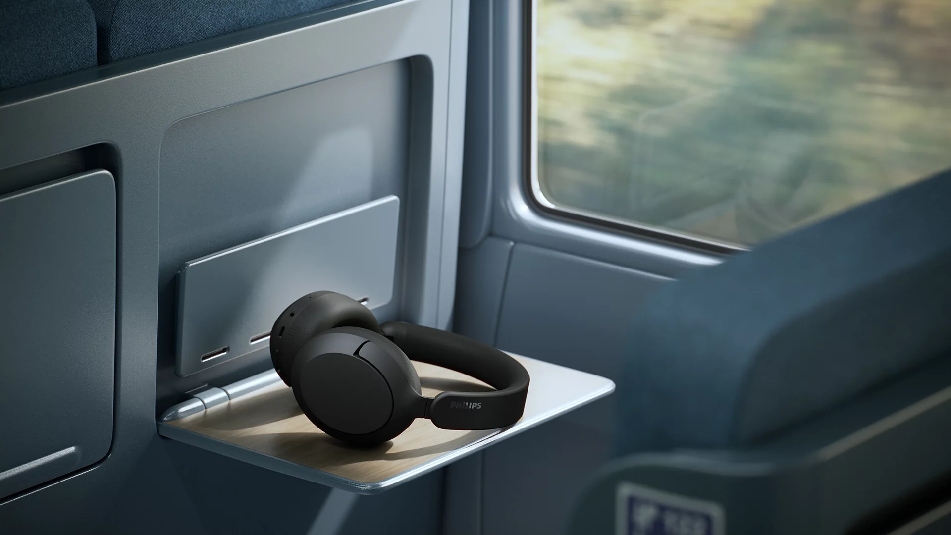 Siyah Philips kulaklık, bir tren masasında pencerenin yanında duruyor.