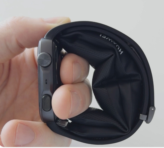 Ręka trzyma czarny smartwatch ze złożonym paskiem na jasnym tle.