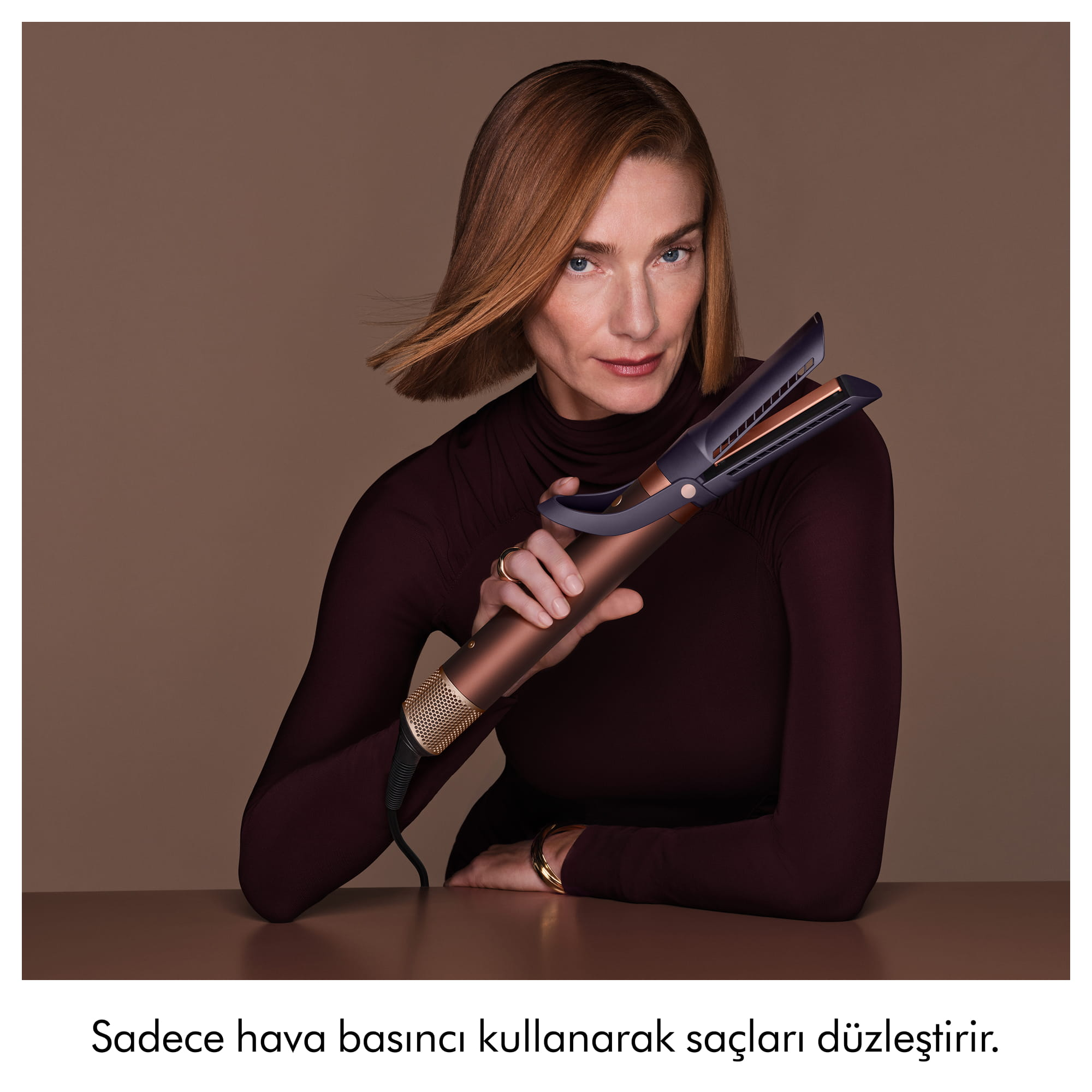 Kahverengi üst giyen bir kadın, düz bir arka plan önünde yenilikçi bir saç şekillendirme aleti tutuyor.