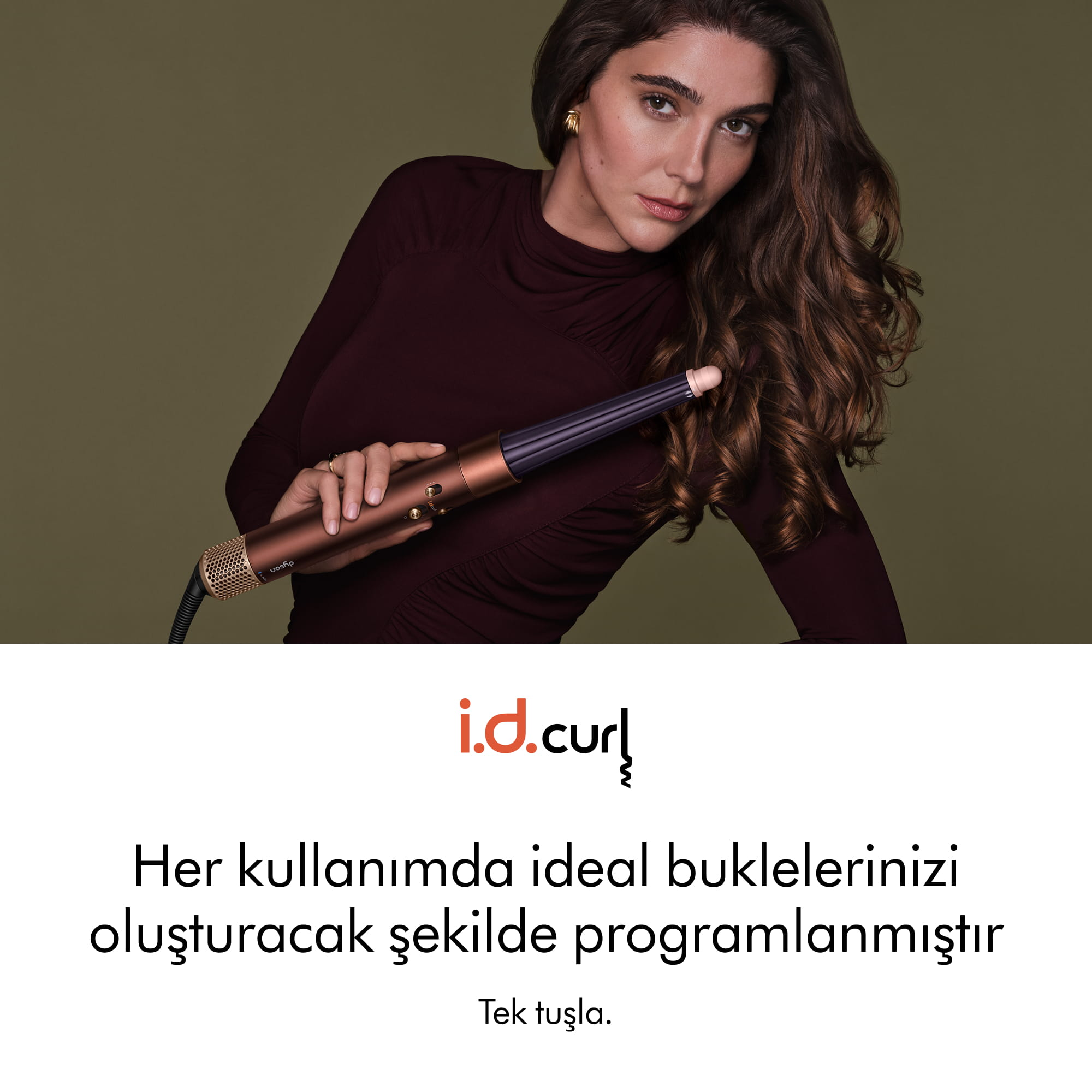 Bordo kazaklı bir kadın, zeytin yeşili arka planda altın rengi bir Dyson maşa tutuyor.