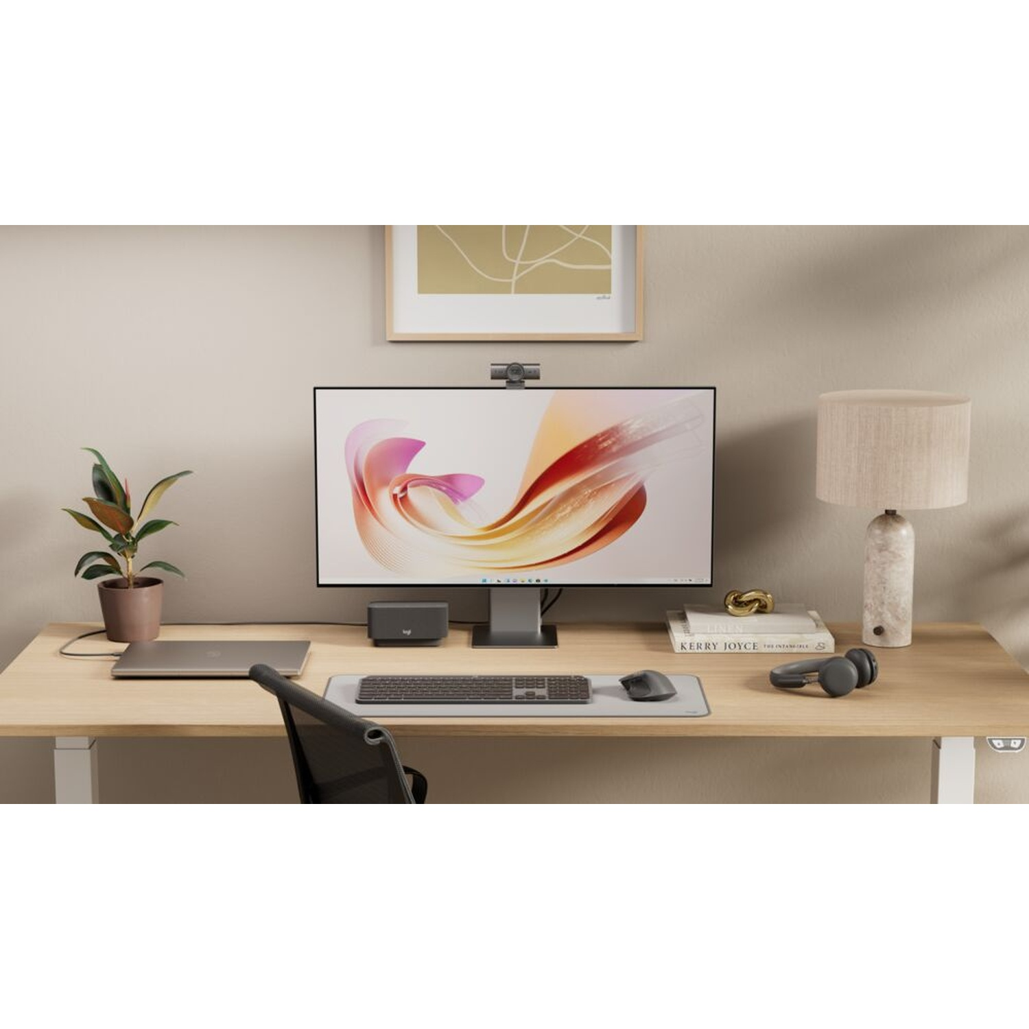 Una scrivania in legno minimalista con monitor, laptop, tastiera, lampada e pianta in vaso contro una parete chiara.