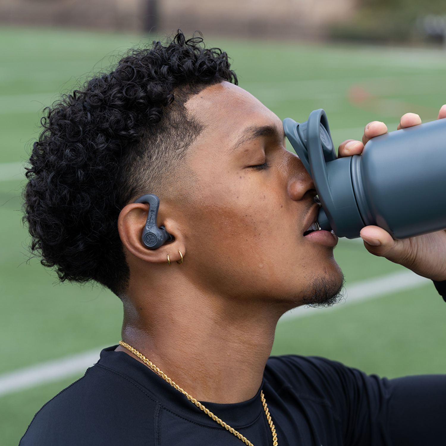 A man with earphones drinks from a blue bottle on a sports field.