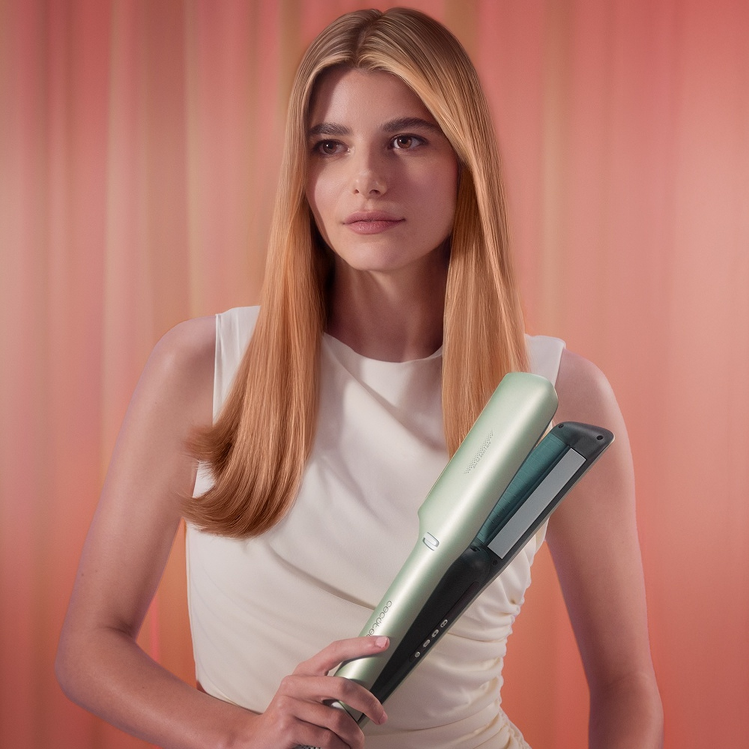 Una donna bionda in bianco tiene una piastra per capelli verde menta davanti a una tenda rosa.