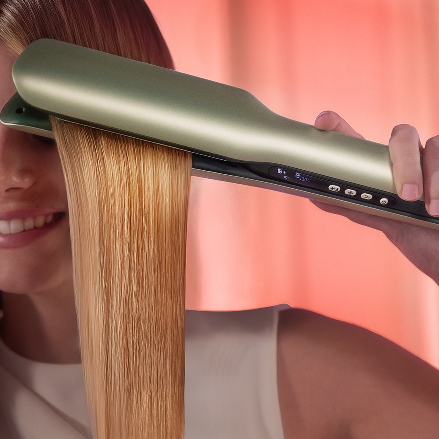 Una donna sorridente liscia i suoi capelli biondi con una moderna piastra verde su sfondo rosa.