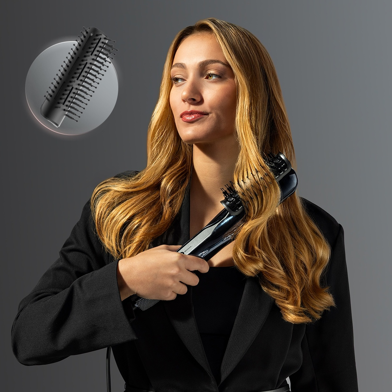 Donna con lunghi capelli biondi e blazer nero acconcia i capelli con una spazzola per capelli su sfondo grigio.