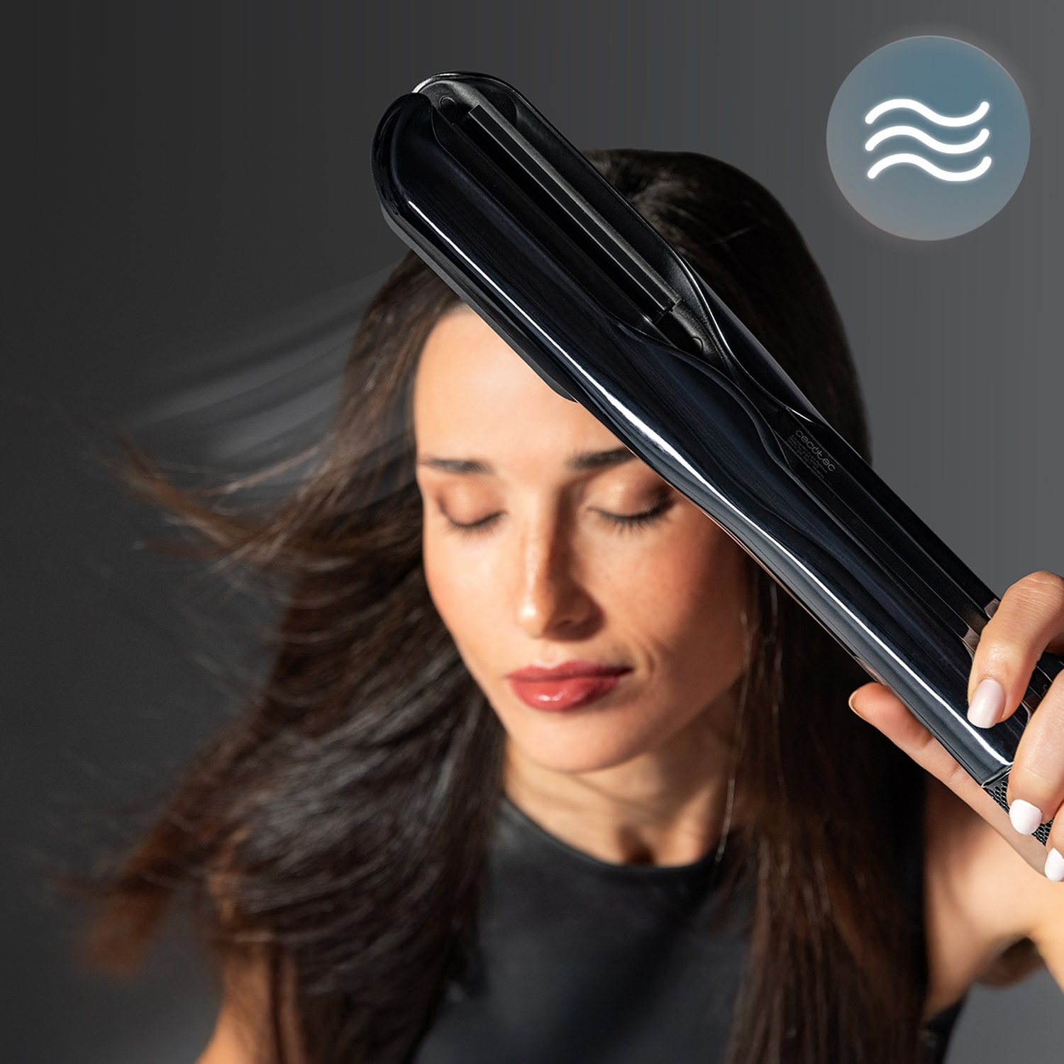 Una donna tiene una piastra per capelli nera sui suoi lunghi capelli castani su sfondo grigio.