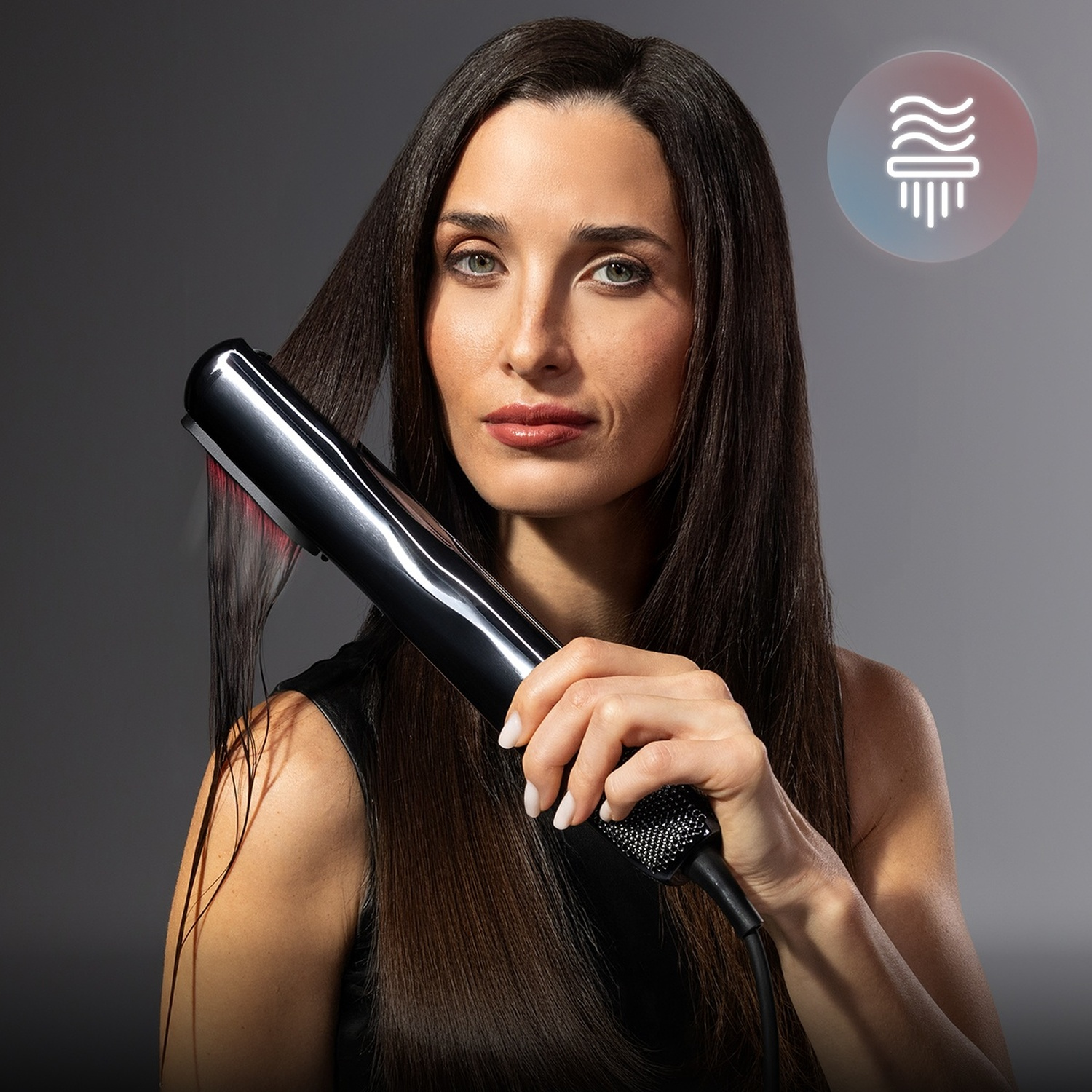 Una donna con lunghi capelli scuri acconcia i capelli usando una piastra nera su sfondo grigio.