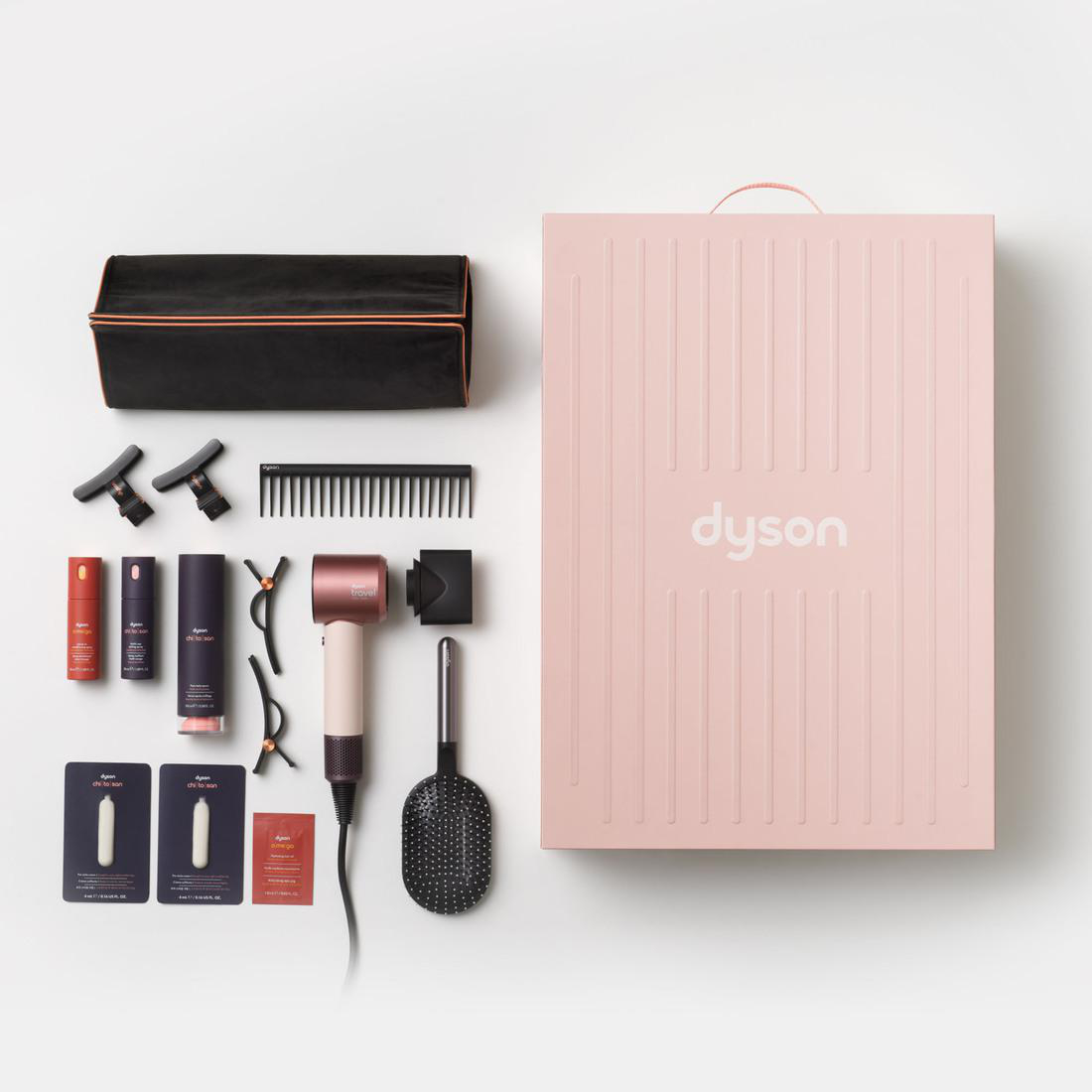 Ein Dyson-Haartrockner mit Zubehör, Bürsten und Produkten neben einer großen rosa Schachtel.