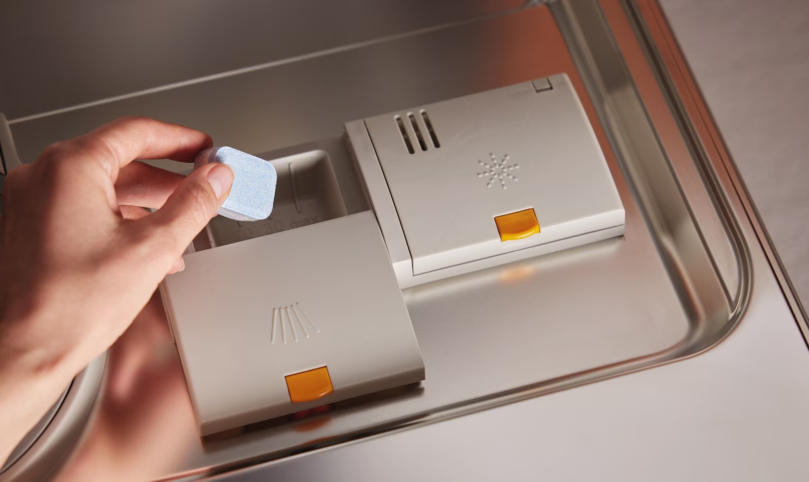 Ręka umieszcza niebieską tabletkę do zmywarki w otwartej przegródce zmywarki.