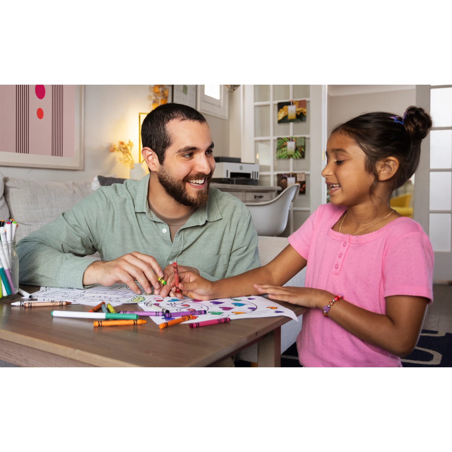A father and daughter sit at a table drawing together with many colorful crayons.