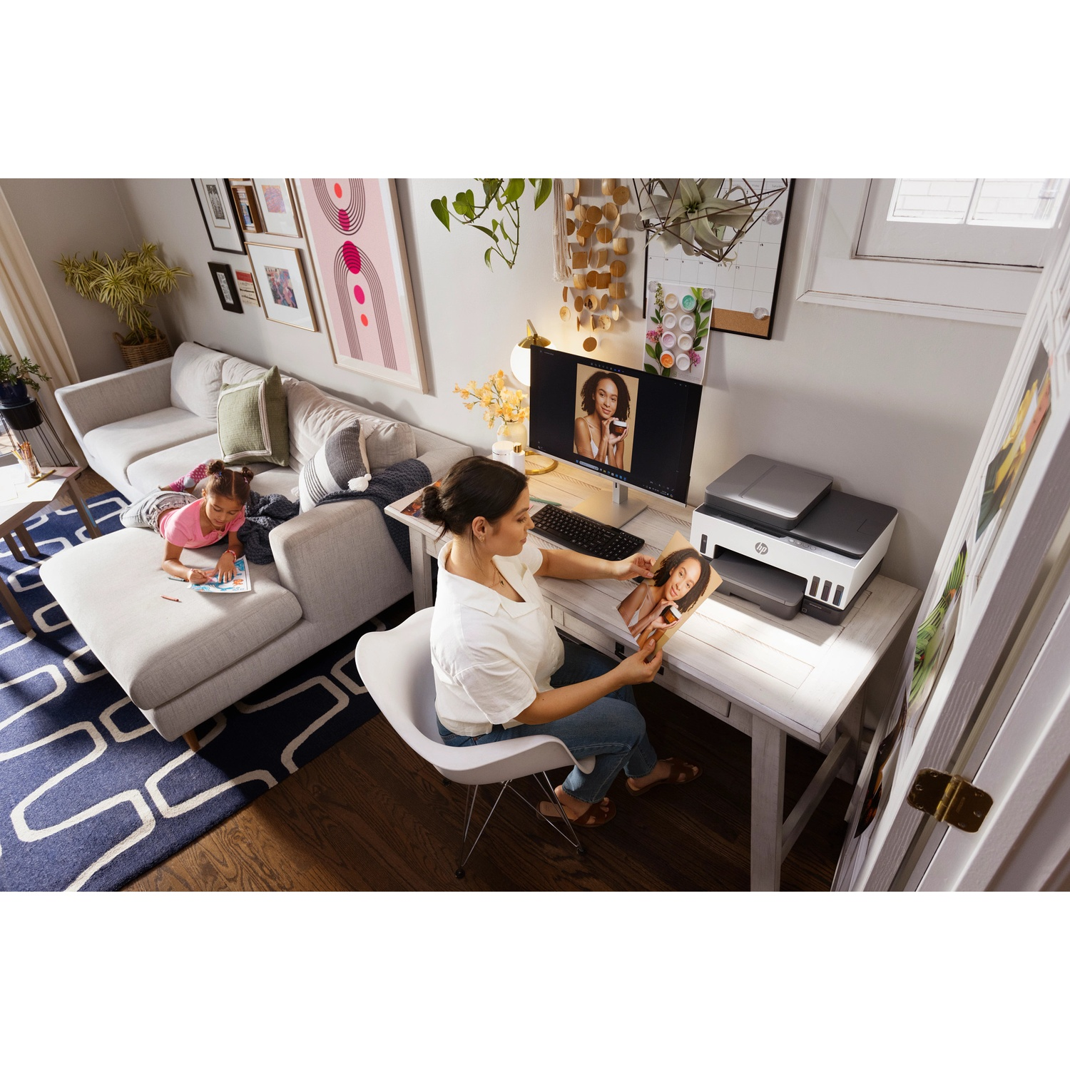 A woman works at a computer in a bright living room while a child draws on the sofa.