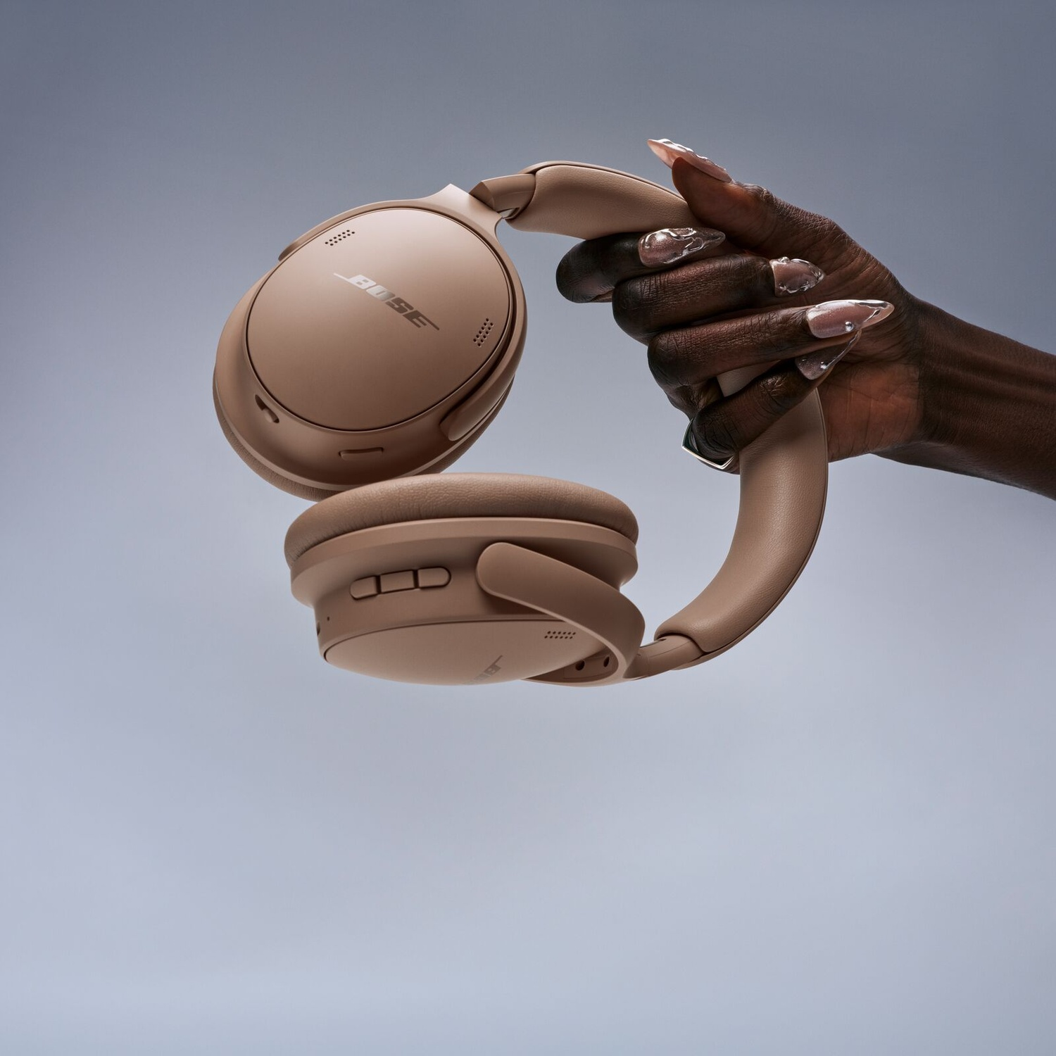 A dark-skinned hand holds beige Bose headphones against a plain gray background.
