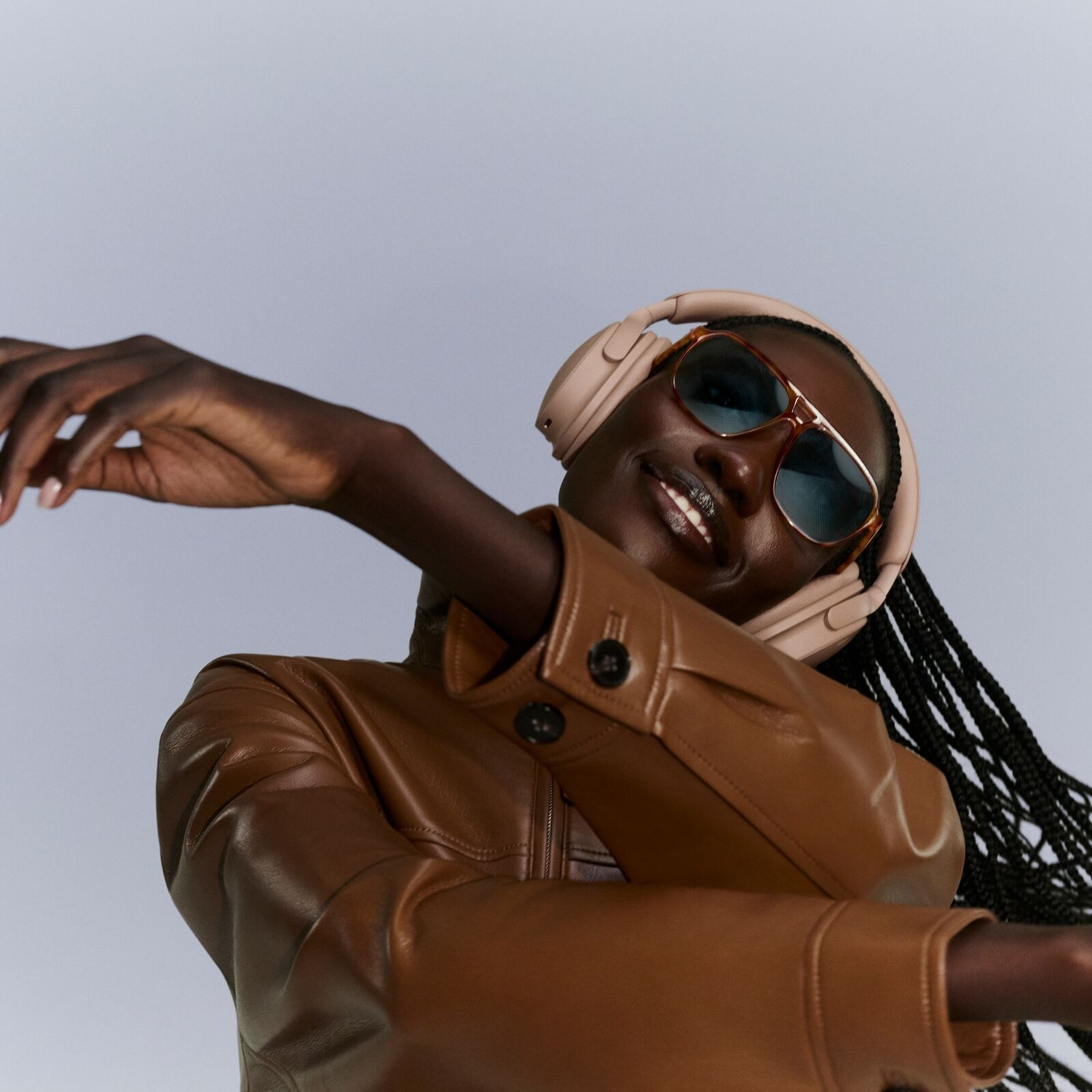 A woman wearing a brown leather jacket, sunglasses, and headphones poses smilingly against a light background.