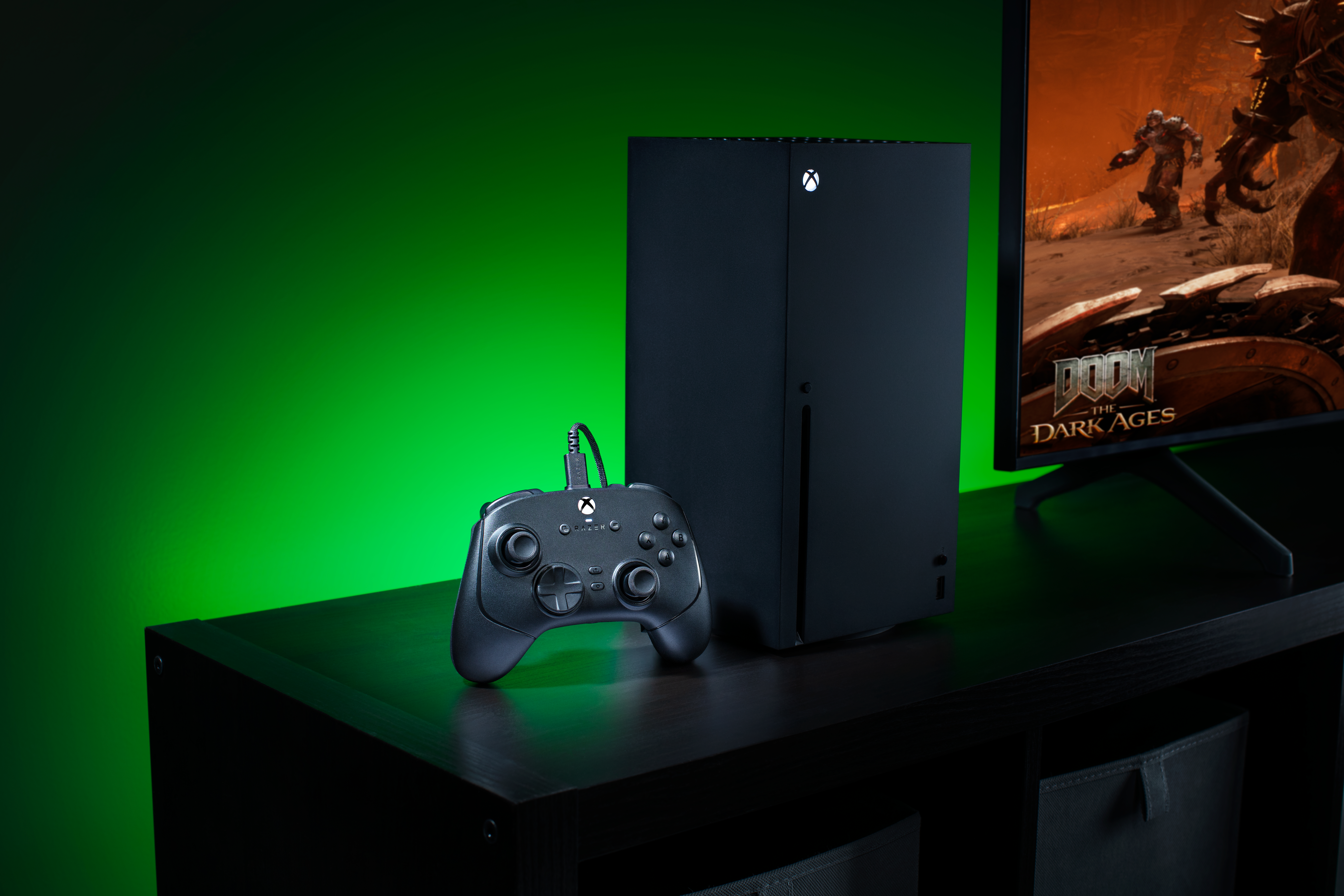 Schwarze Xbox Konsole und Controller auf einem Tisch vor grünem Hintergrund und Bildschirm.