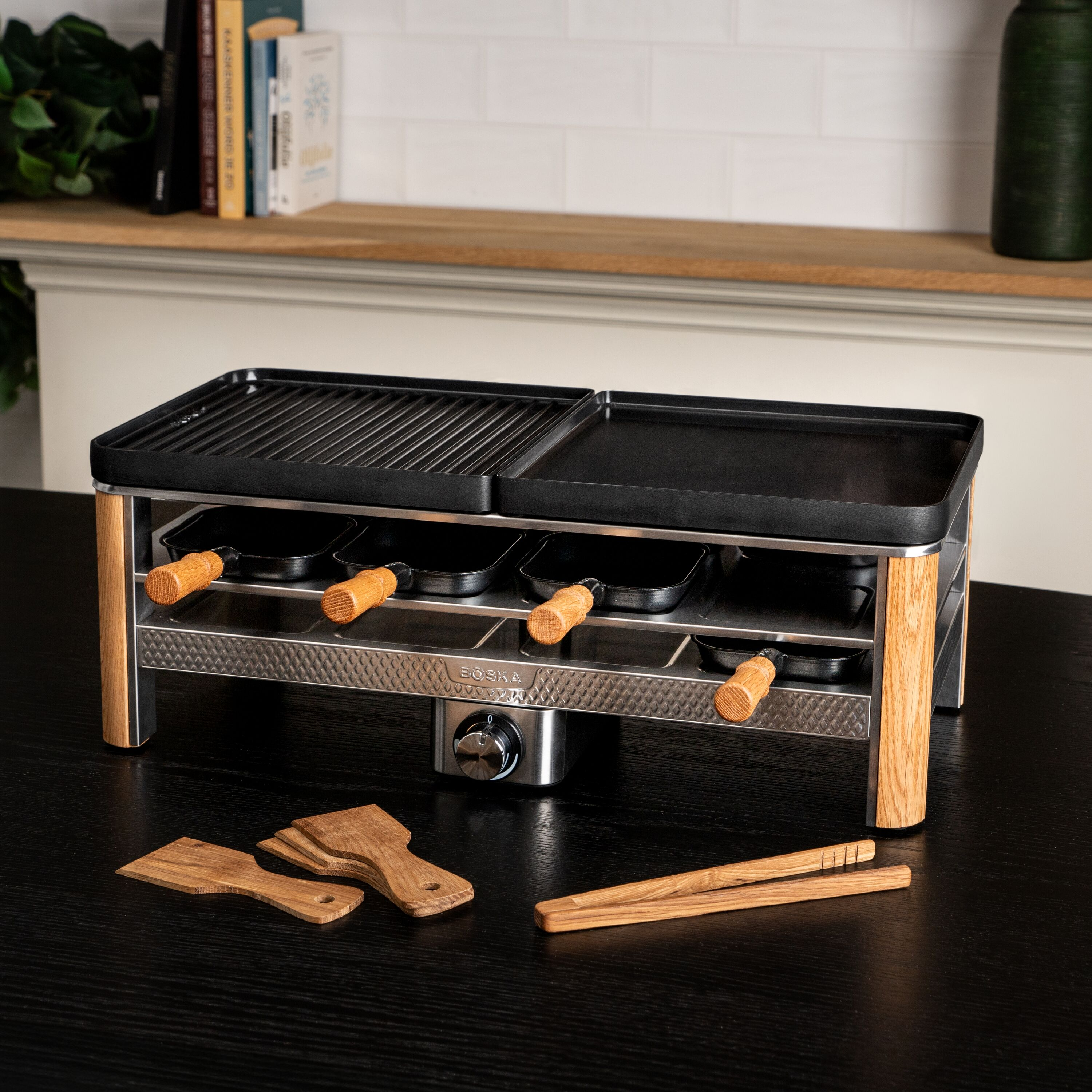 Ein schwarzer Raclette-Grill mit Holzakzenten steht auf einem dunklen Tisch, mit Spateln und Zange vorne.