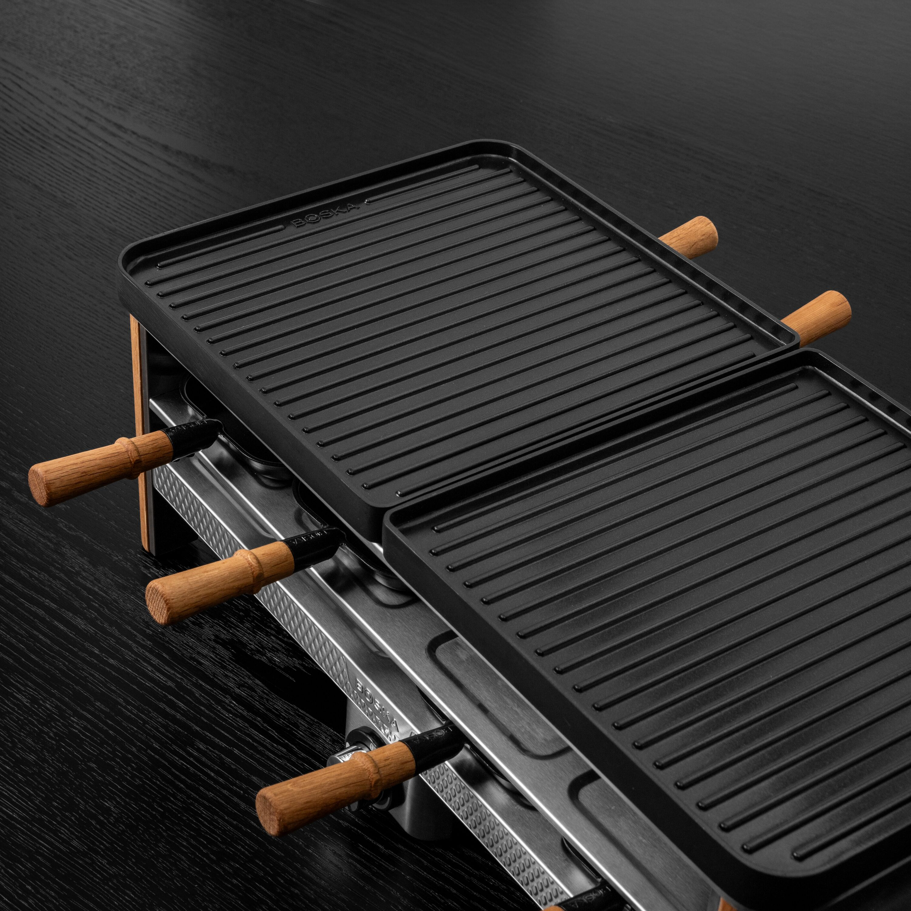 Schwarzer Raclette-Grill mit Holzgriffen auf dunklem Holztisch.