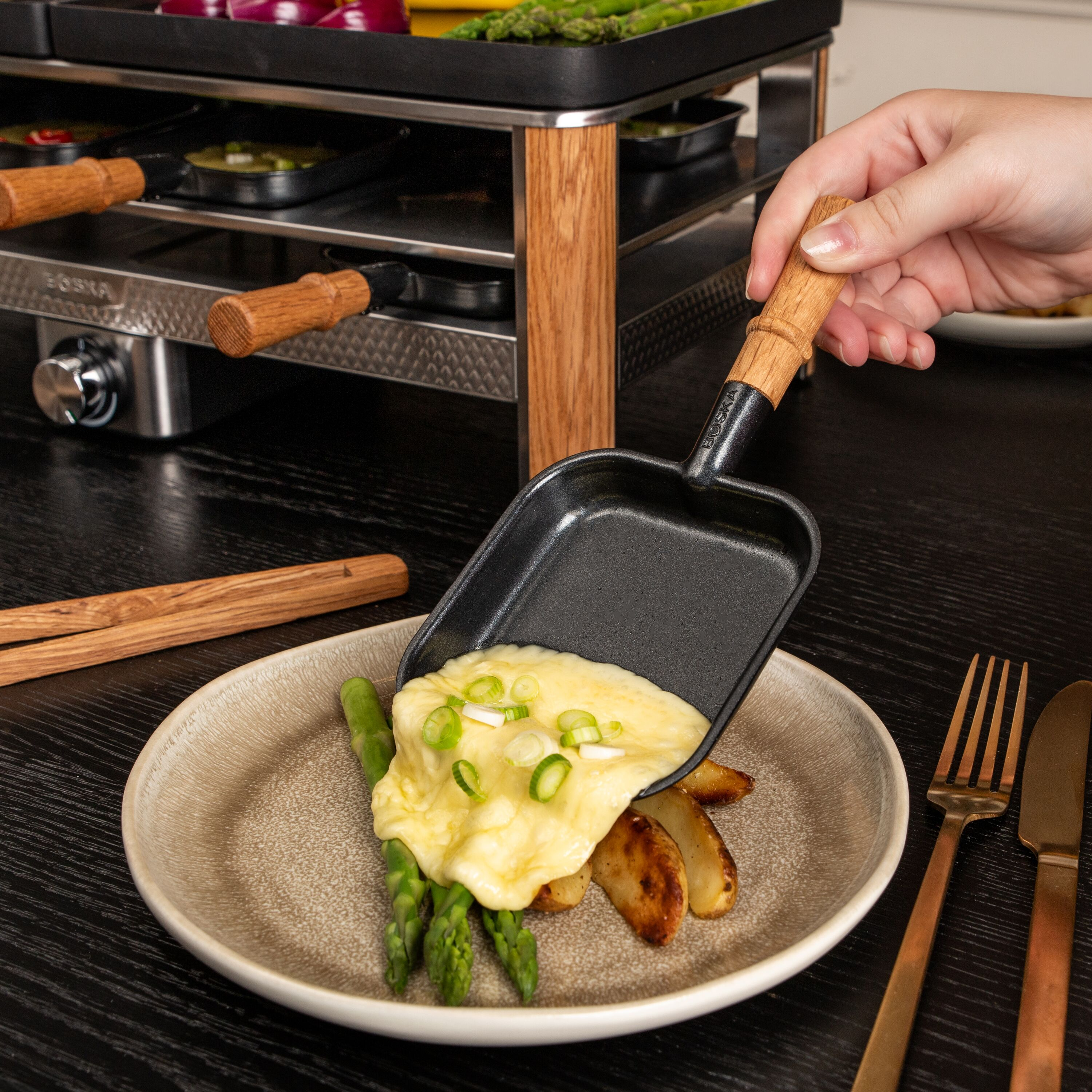 Hand gießt geschmolzenen Käse von einer Raclettepfanne auf Spargel und Kartoffeln.