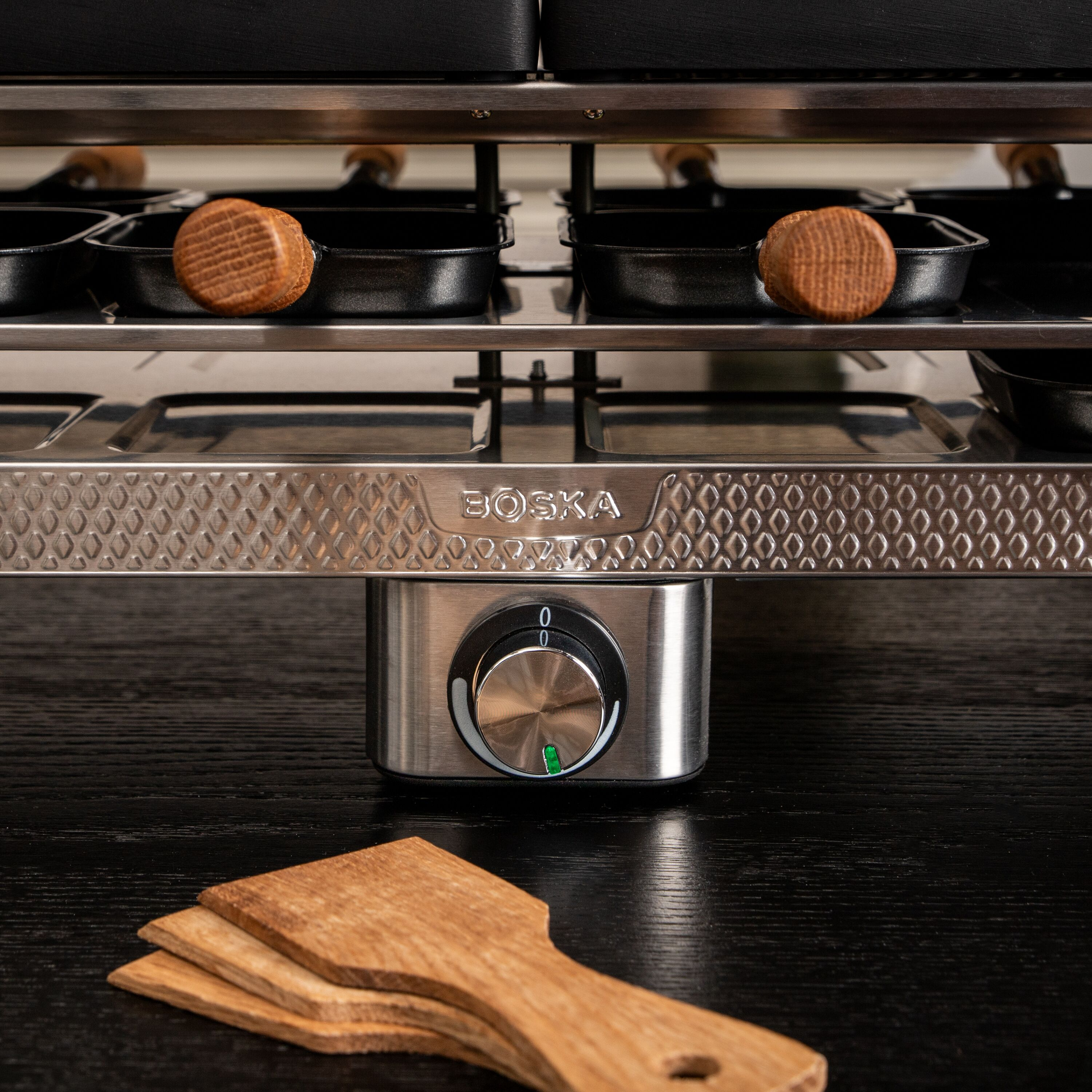 Bosca Raclette-Grill aus Edelstahl mit schwarzen Pfännchen, Holzzylindern und kleinen Holzschaufeln.