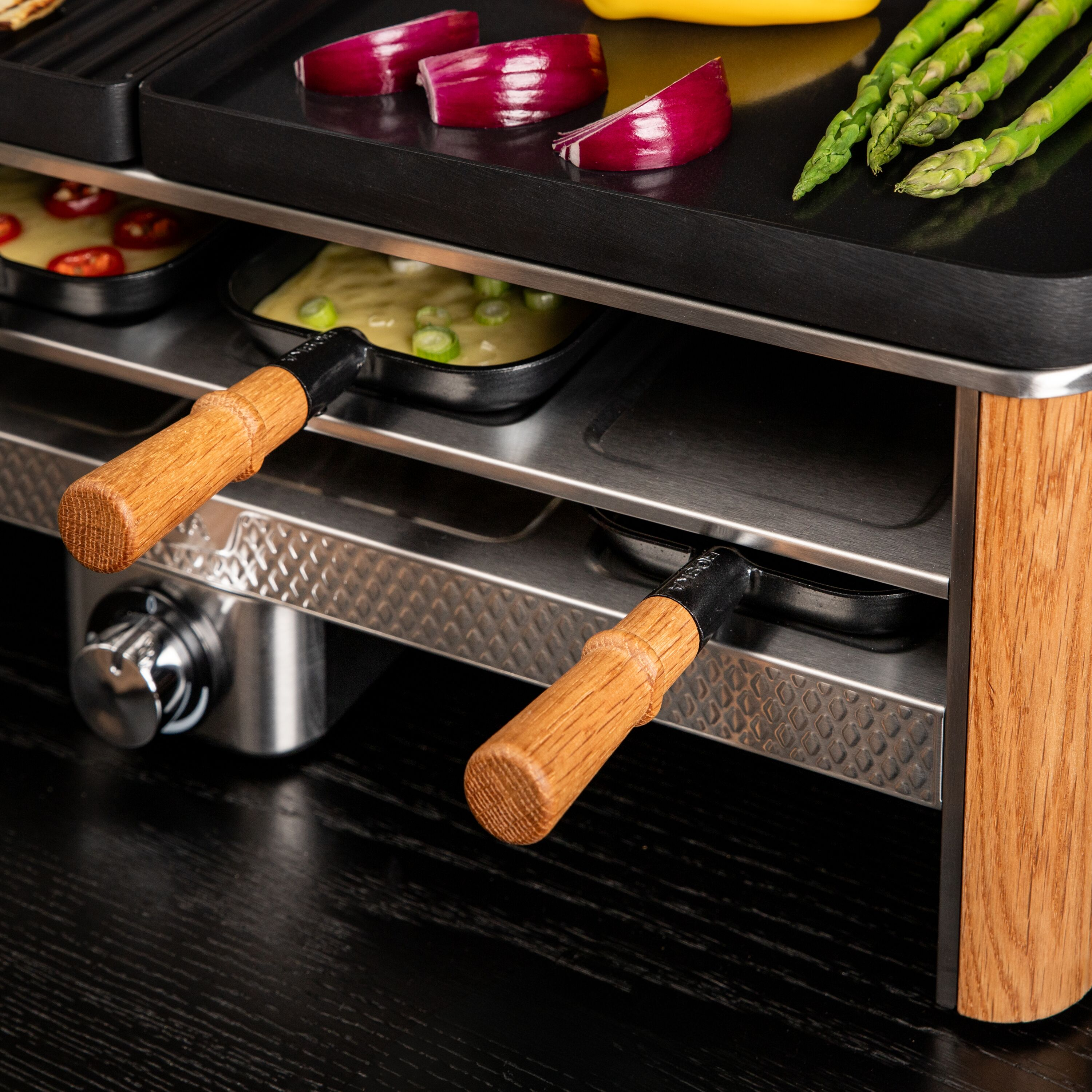 Ein Raclette-Grill mit Holzdetails steht auf einem Tisch, oben mit Zwiebeln und Spargel, unten mit Pfännchen.