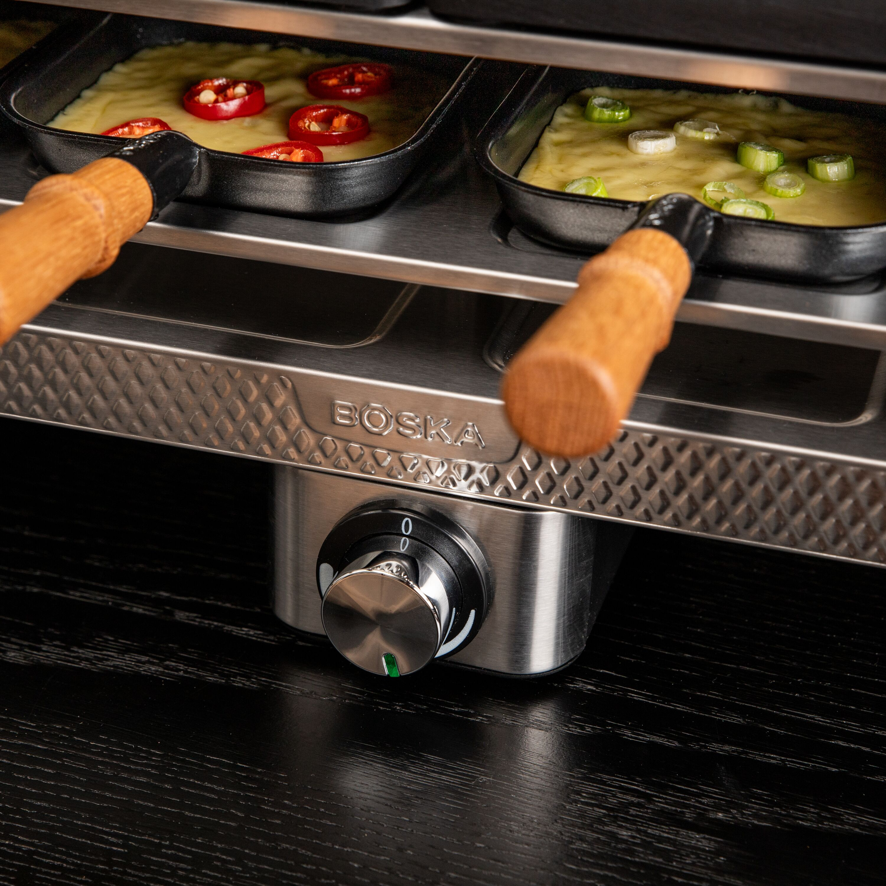 Edelstahl-Raclette-Grill von Boska mit Pfännchen, die mit geschmolzenem Käse und Gemüse gefüllt sind.