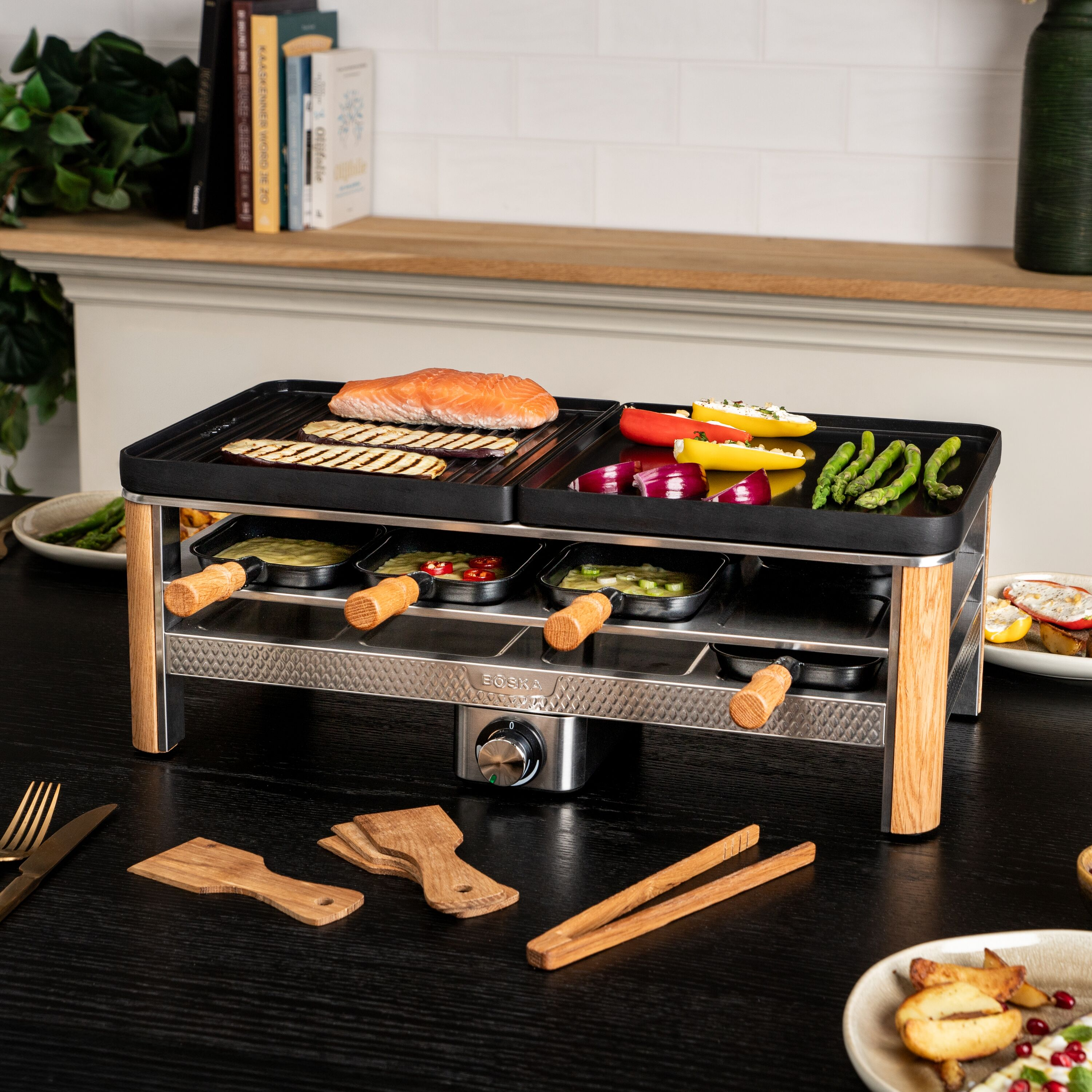 Ein elektrischer Raclette-Grill auf einem Holztisch mit Lachs, Gemüse und kleinen Pfännchen.