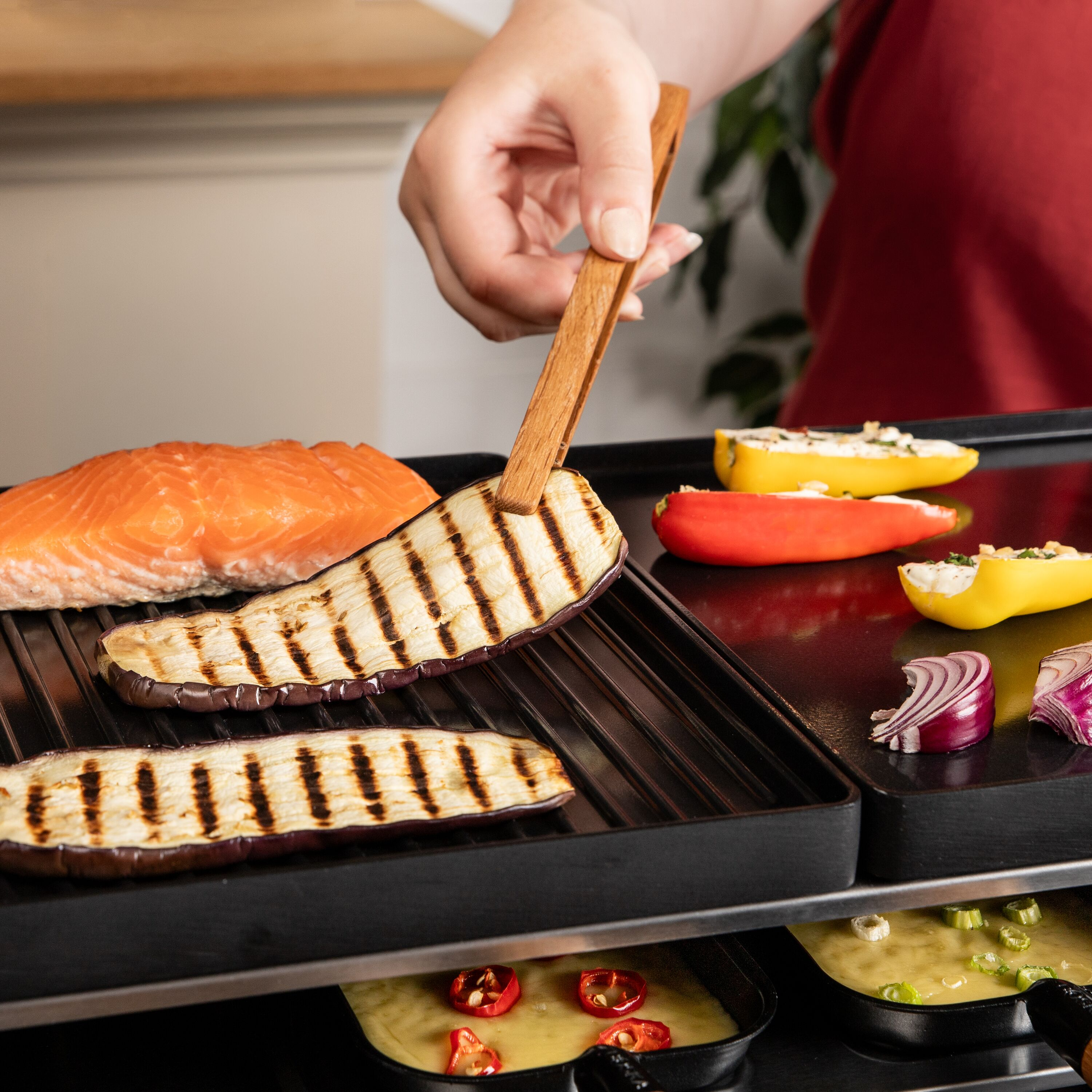 Hand greift mit Holzpinzette eine gegrillte Auberginenscheibe auf einem Tischgrill neben Lachs und Gemüse.