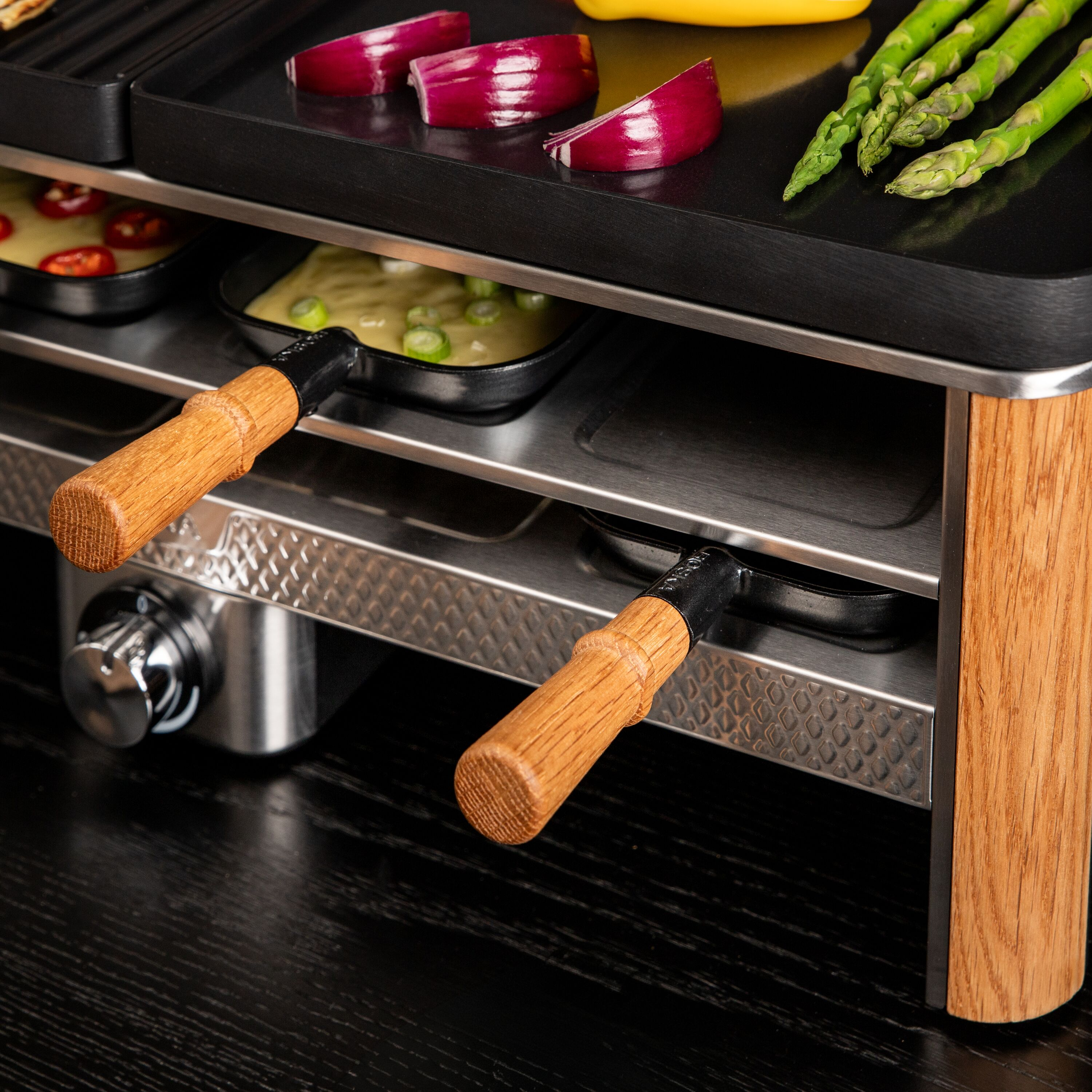 Ein Raclette-Grill mit Holzdetails kocht Zwiebeln, Spargel und kleine Pfännchen auf einem Tisch.