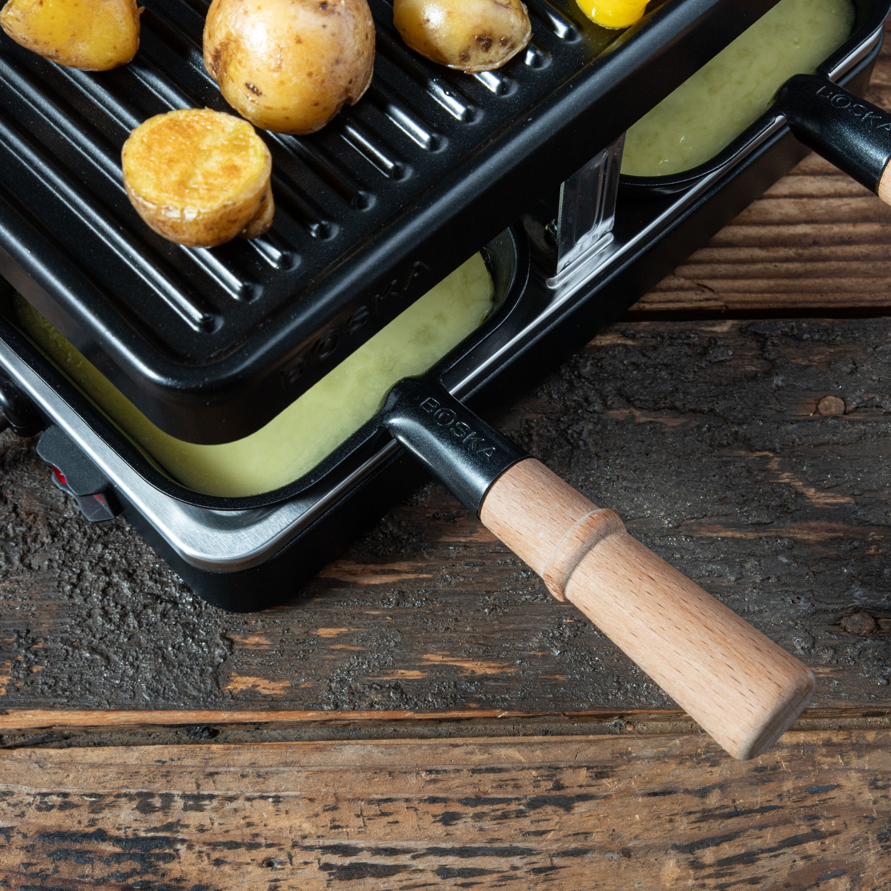 Ein Raclette-Grill mit geschmolzenem Käse in Pfännchen und Kartoffeln oben auf dem Rost, auf Holzhintergrund.