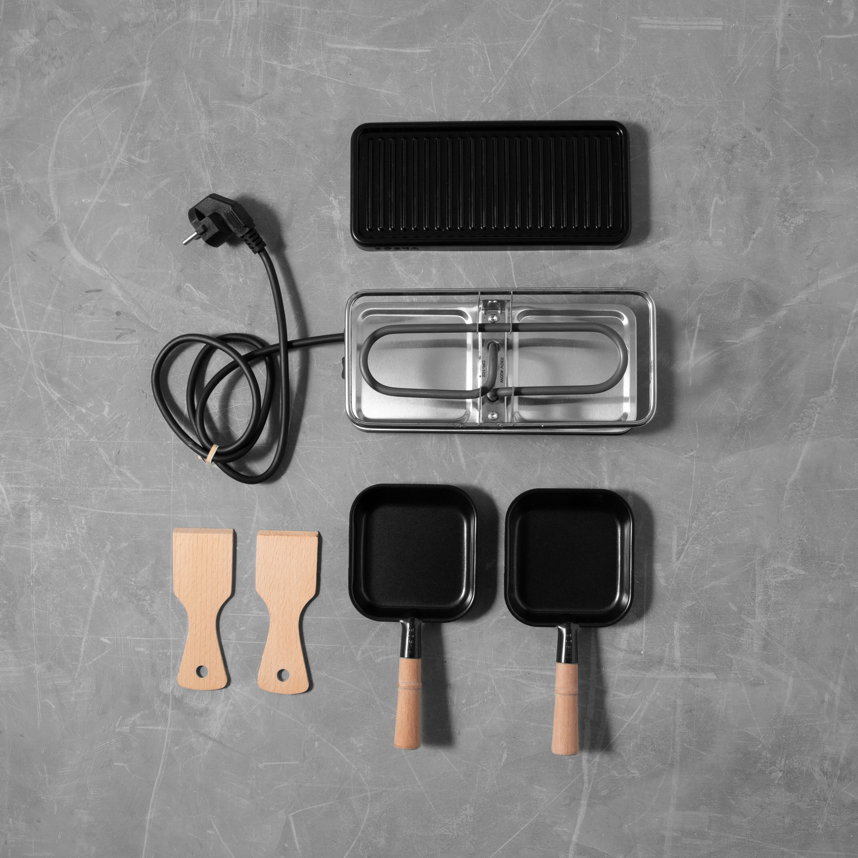 Raclette-Grill-Set mit Heizbasis, Grillplatte, zwei kleinen Pfännchen und zwei Holzspateln auf grauem Stein.