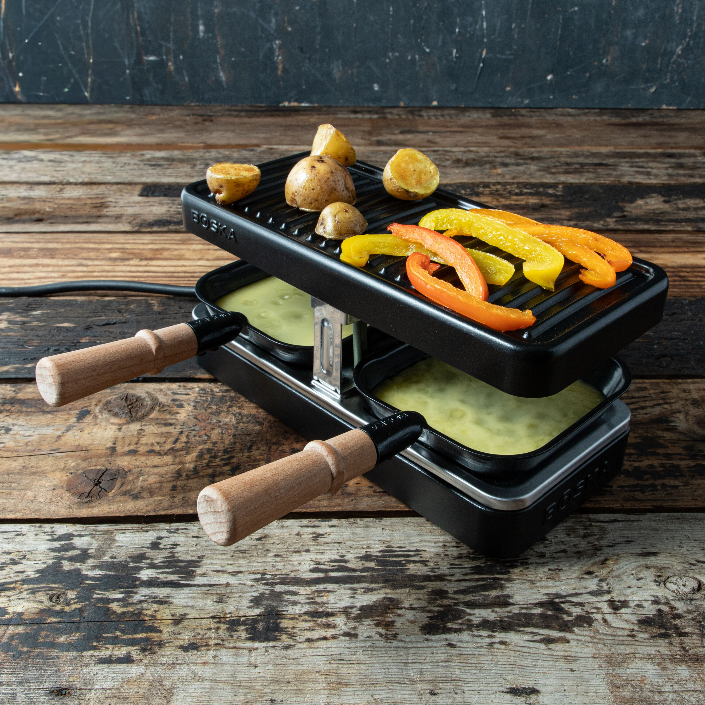 Ein schwarzer Tisch-Raclette-Grill mit Kartoffeln und Paprikastreifen oben, auf einem Holztisch.