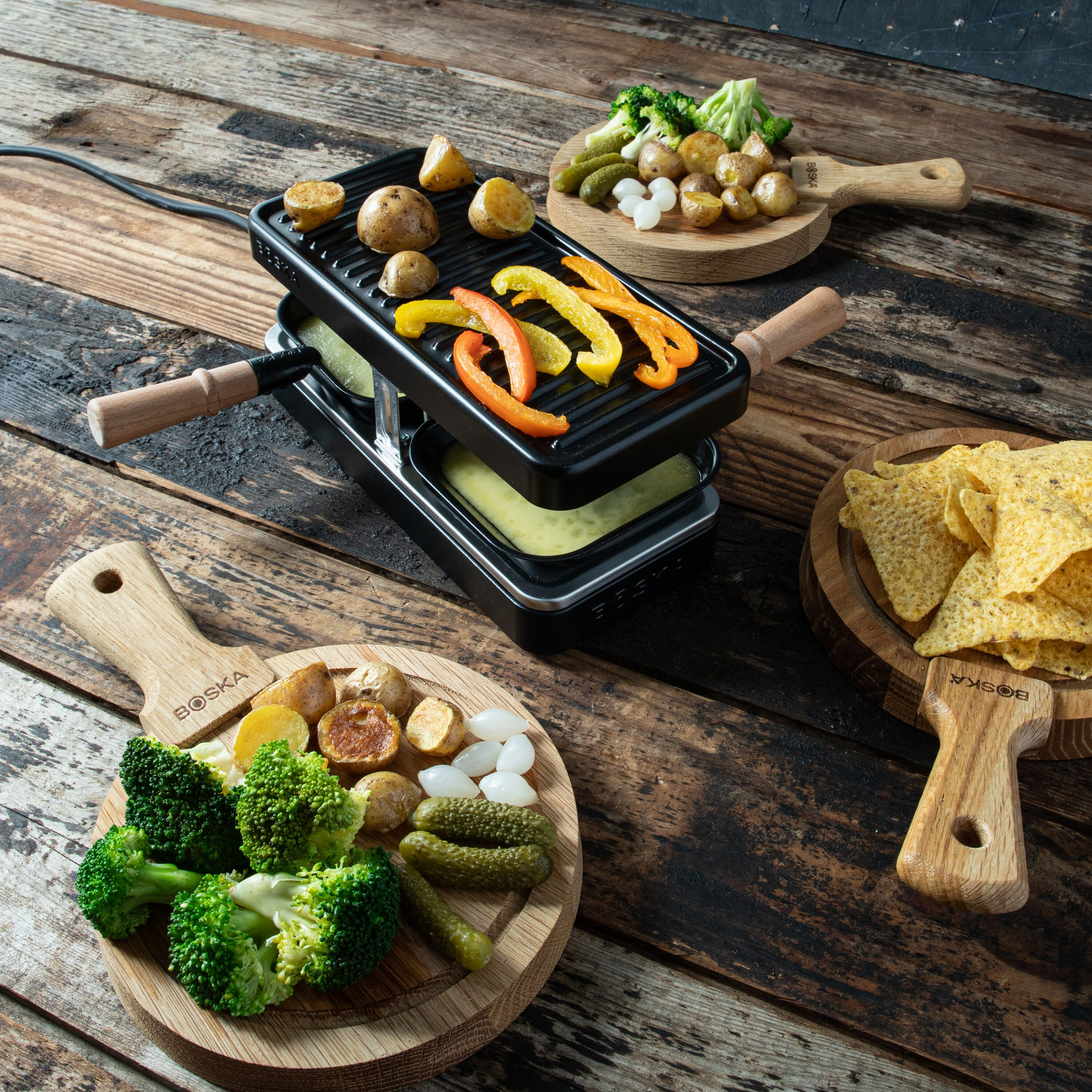 Ein Raclette-Gerät auf einem Holztisch, umgeben von Holzbrettern mit Brokkoli, Kartoffeln, Gurken und Tortilla-Chips.