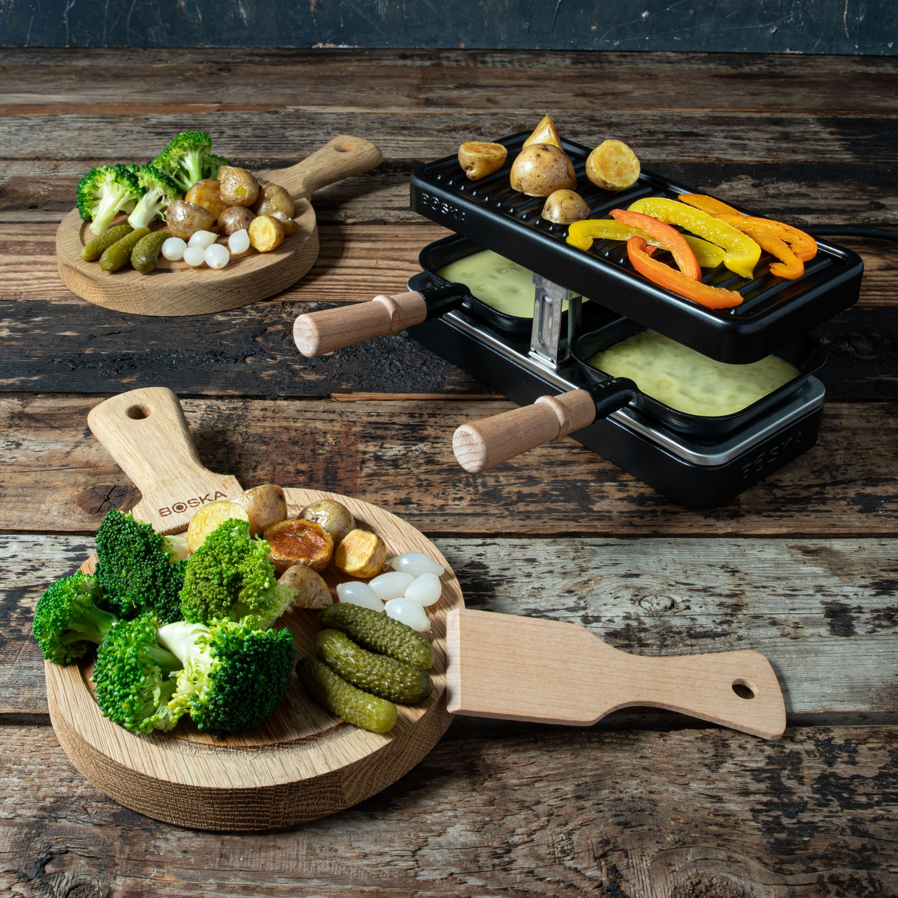 Ein Raclette-Gerät auf Holztisch mit Brokkoli, Kartoffeln und Gurken auf Holzbrettern.