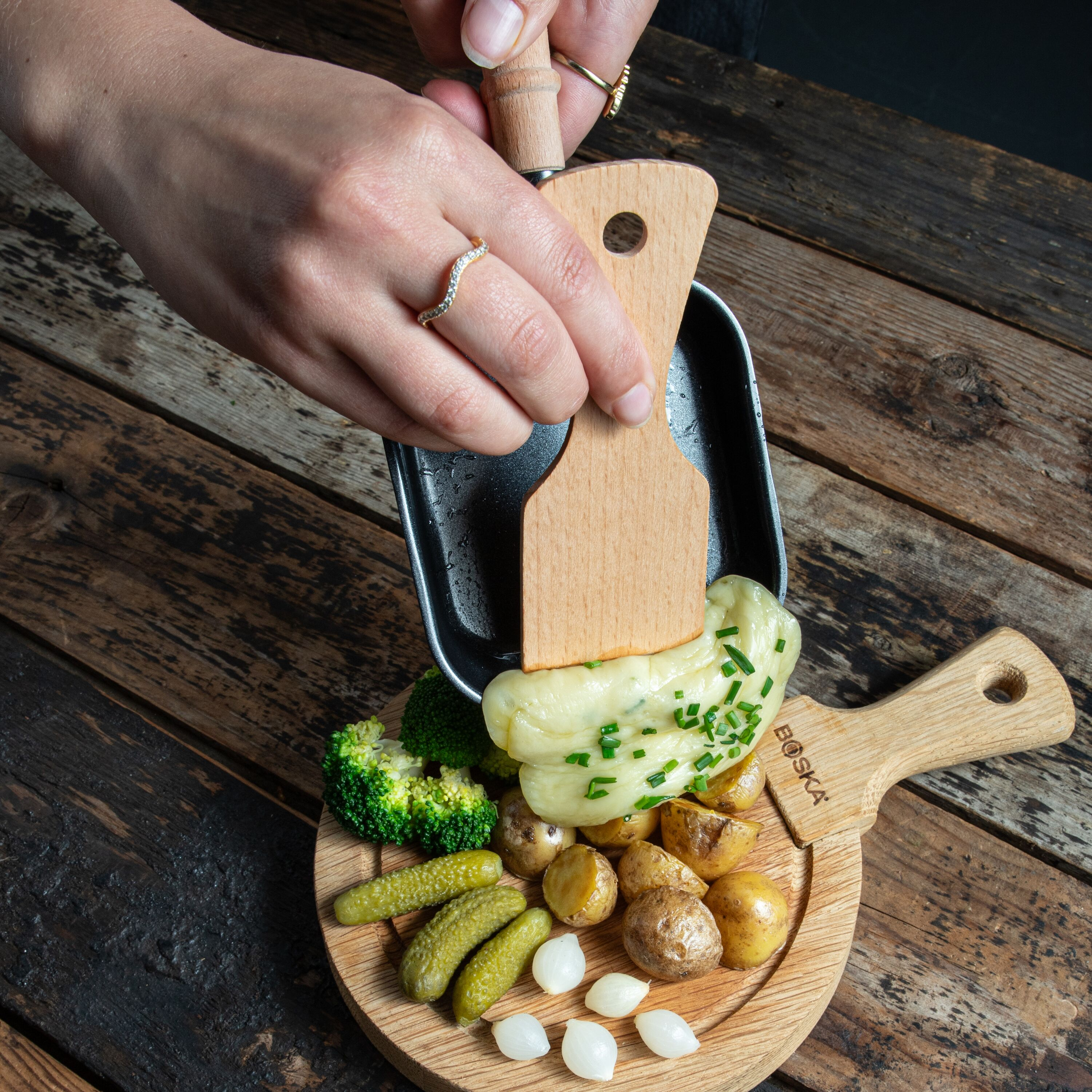Geschmolzener Käse wird mit einem Holzspatel aus einer Pfanne auf Kartoffeln, Brokkoli und Gurken gegossen.