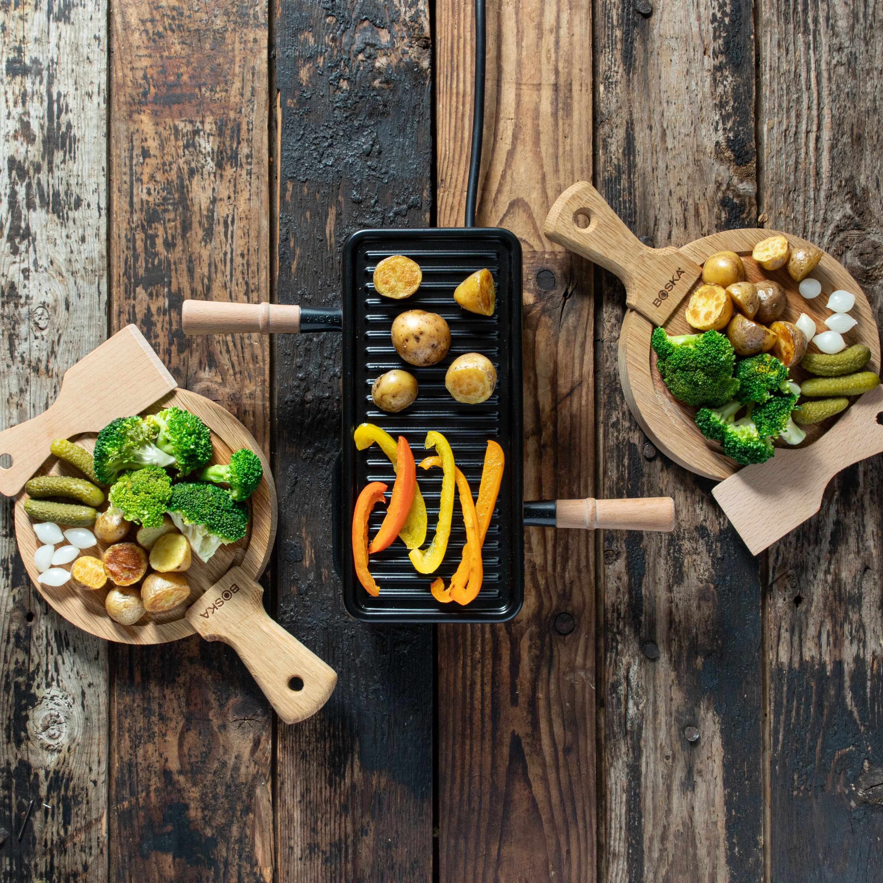 Grillplatte mit Kartoffeln und Paprika, flankiert von Holztellern mit Brokkoli, Pickles und Zwiebeln auf Holz.
