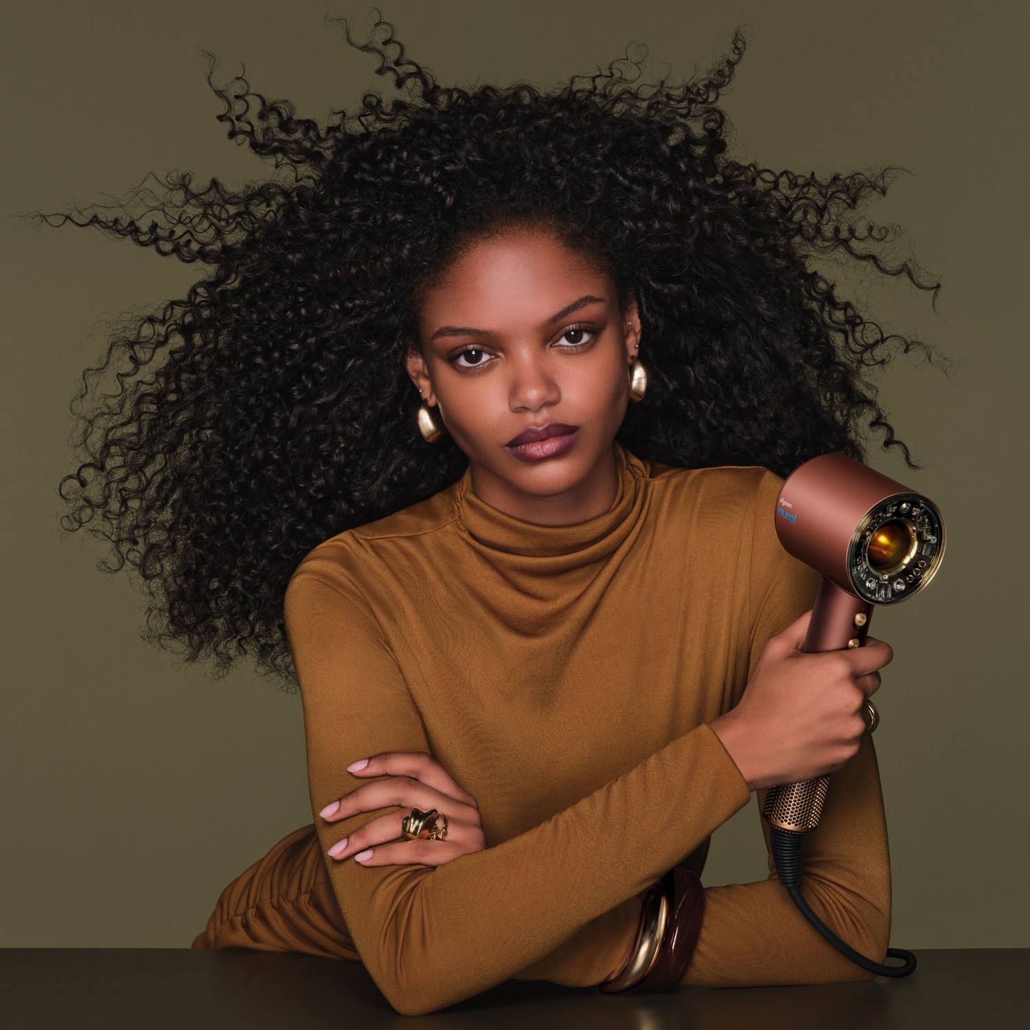 A woman with large curly hair holds a gold hairdryer against an olive green background.
