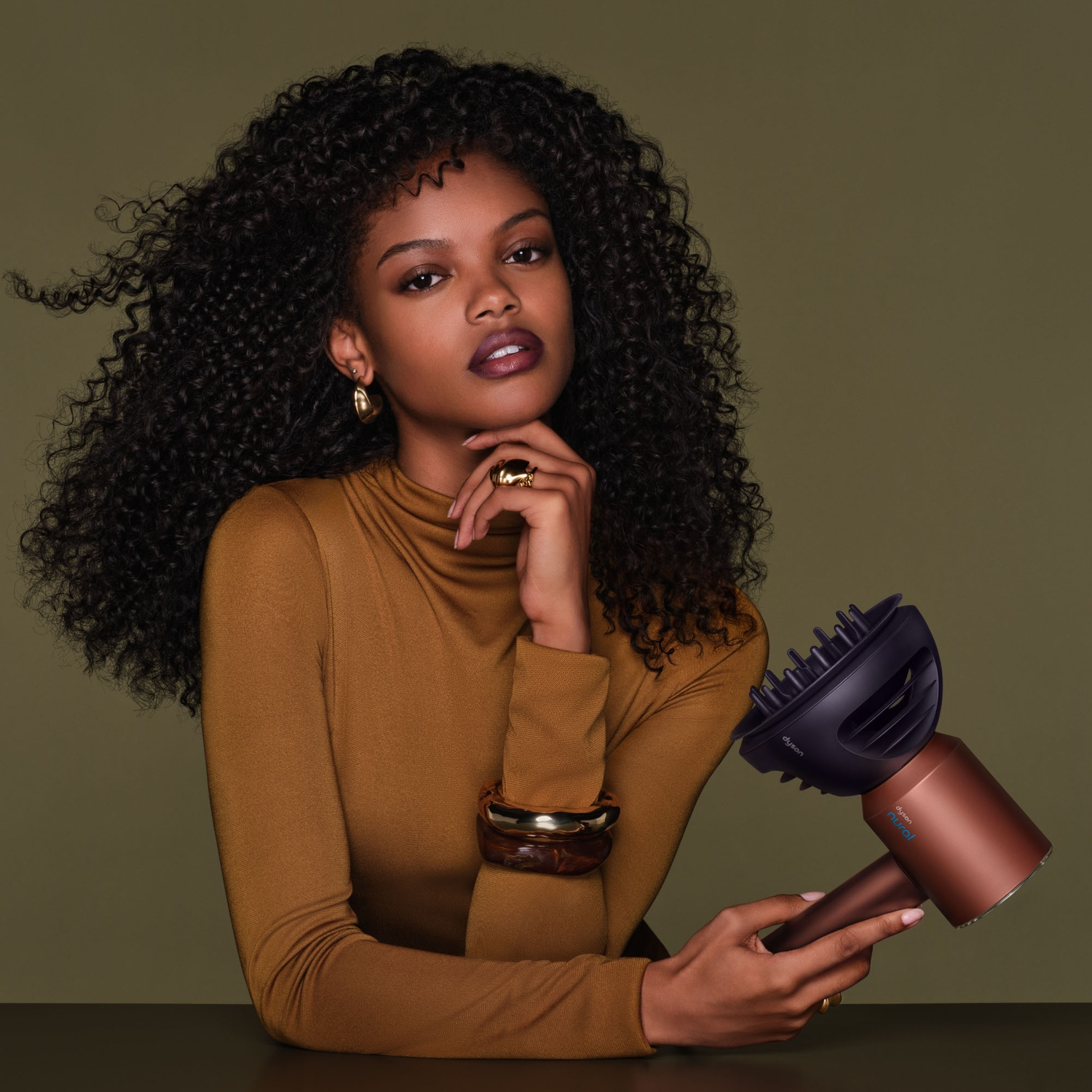 A woman with voluminous curly hair wears a brown top and holds a modern bronze and black hairdryer against a green background.