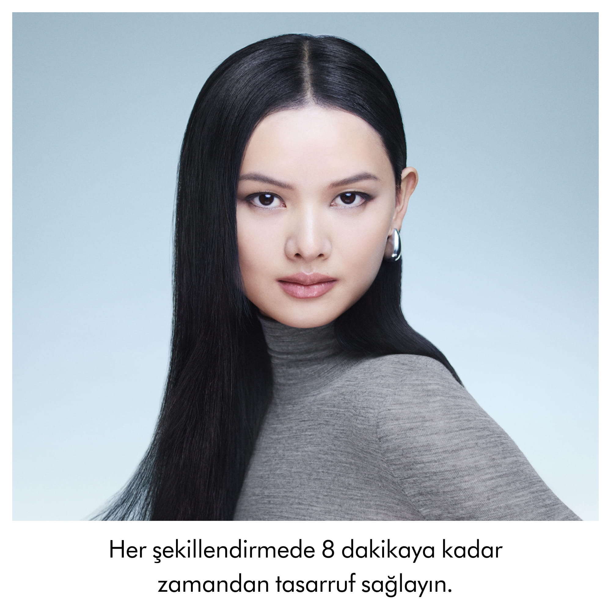Uzun, düz siyah saçlı bir kadın, açık arka plan önünde gri balıkçı yaka kazak giyiyor.