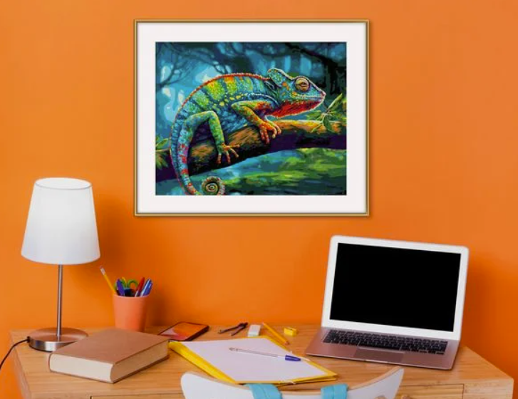 Ein Schreibtisch mit Laptop, Lampe und Notizbuch vor einer orangefarbenen Wand mit Chamäleon-Bild.
