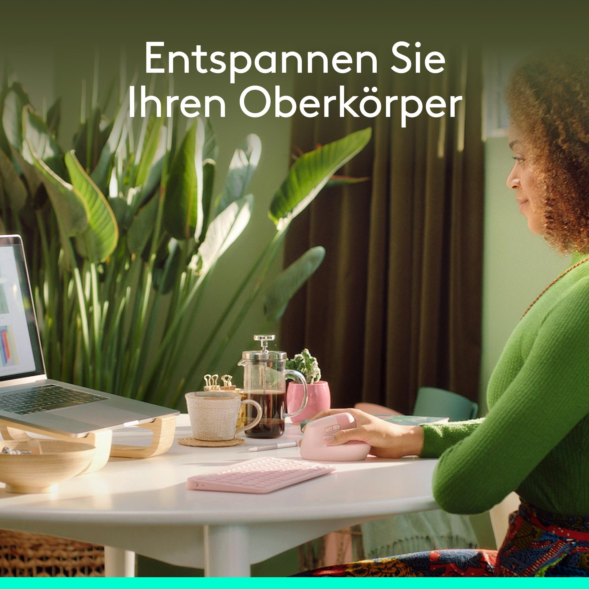 Eine Frau in grünem Pullover arbeitet an einem Laptop an einem weißen Tisch mit Pflanzen im Hintergrund.