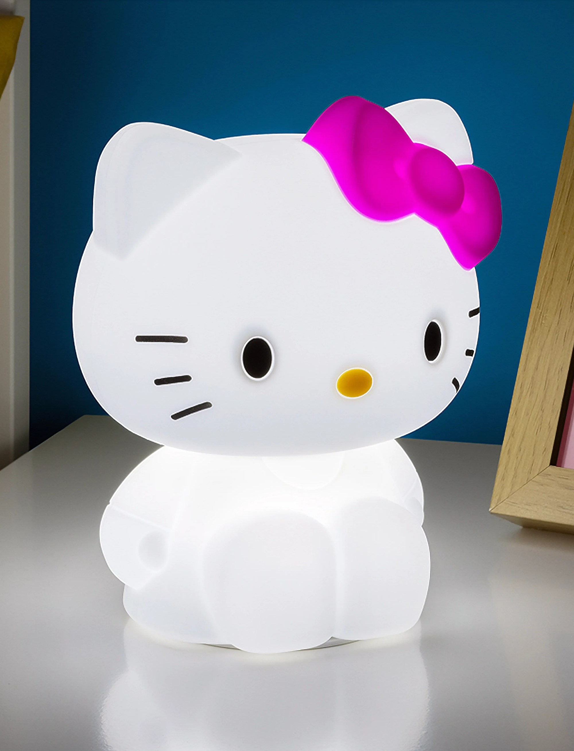 Egy megvilágított fehér Hello Kitty lámpa élénk rózsaszín masnival áll egy fehér asztalon.