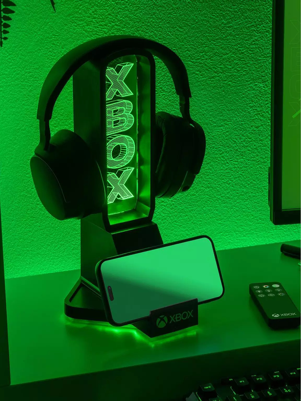 Fekete Xbox fejhallgató-állvány zöld LED logóval, fejhallgatót és okostelefont tart zöld fal előtt.