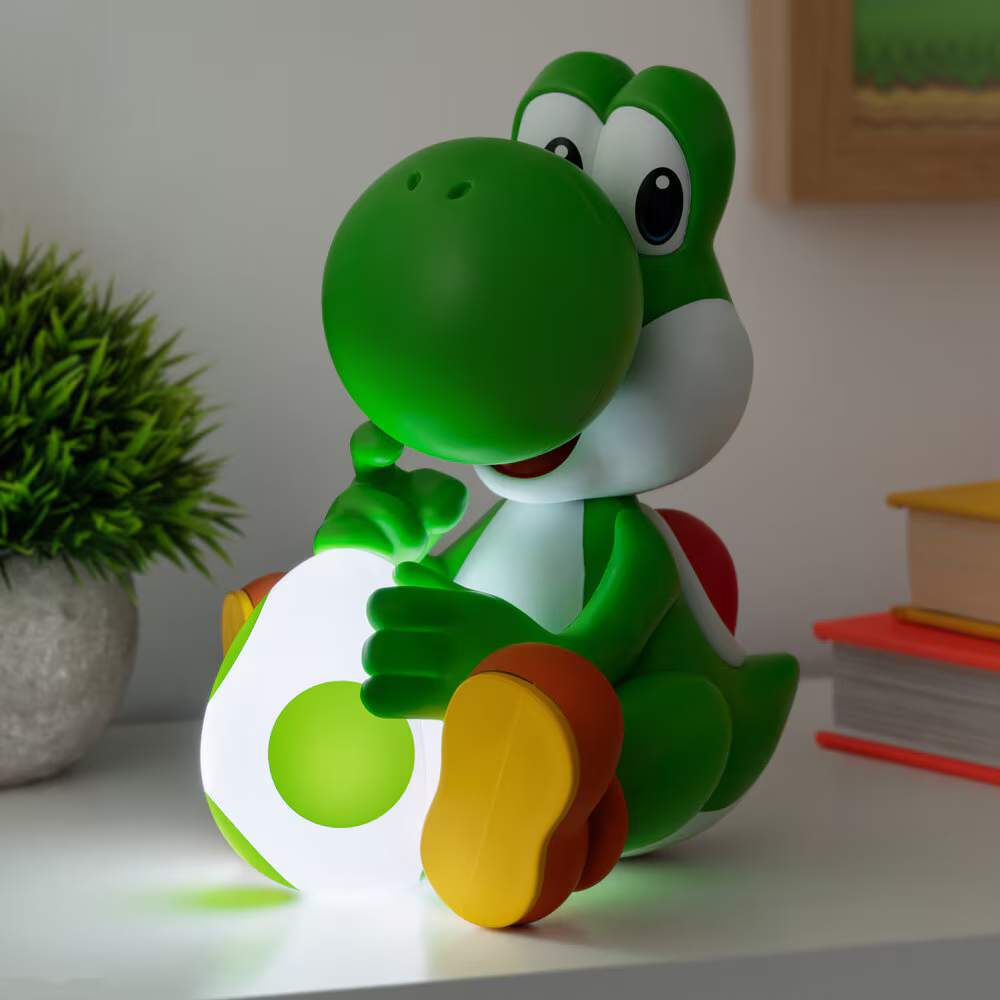 Egy zöld Yoshi figura ül az asztalon, egy világító tojást tartva, könyvekkel és egy növénnyel a háttérben.