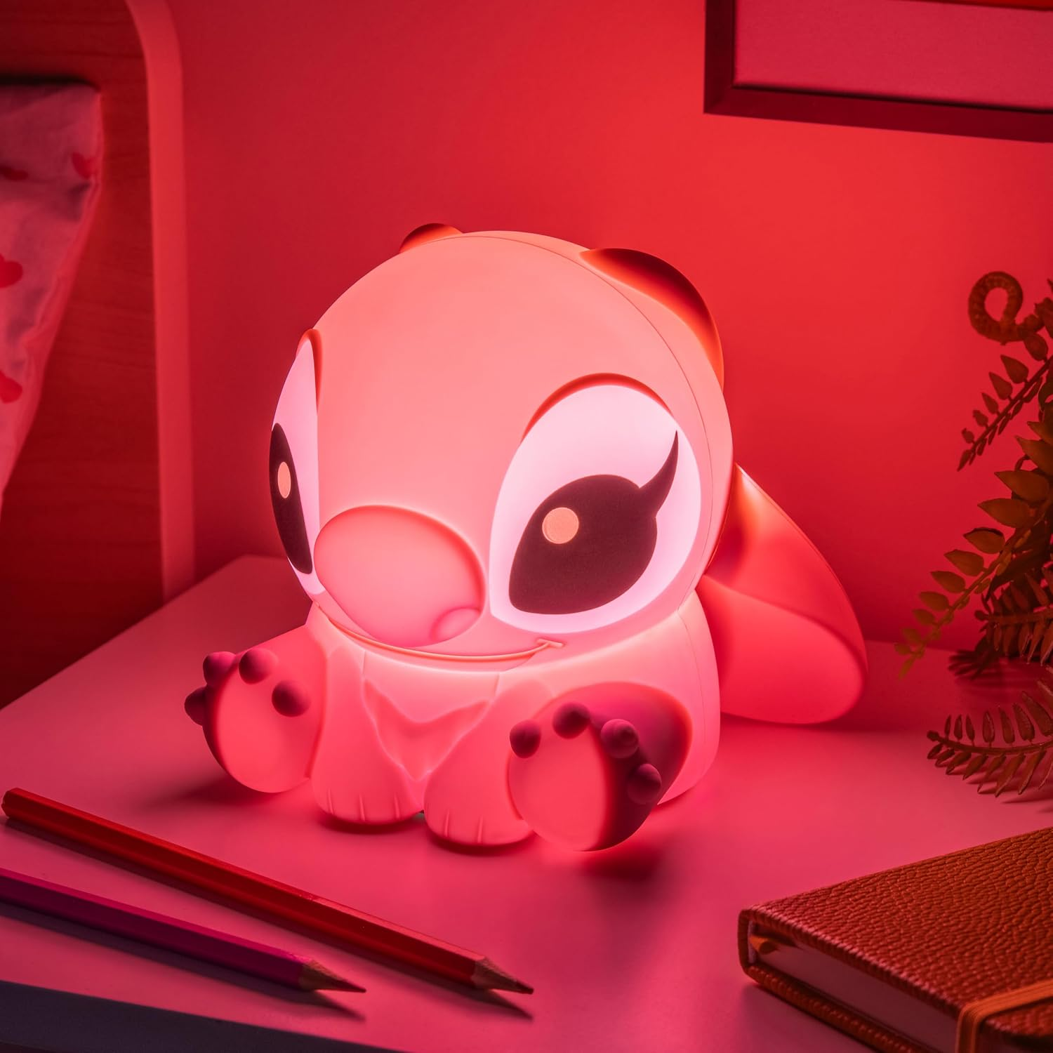 Egy izzó rózsaszín Stitch éjjeli lámpa ül az asztalon ceruzák és egy füzet mellett.