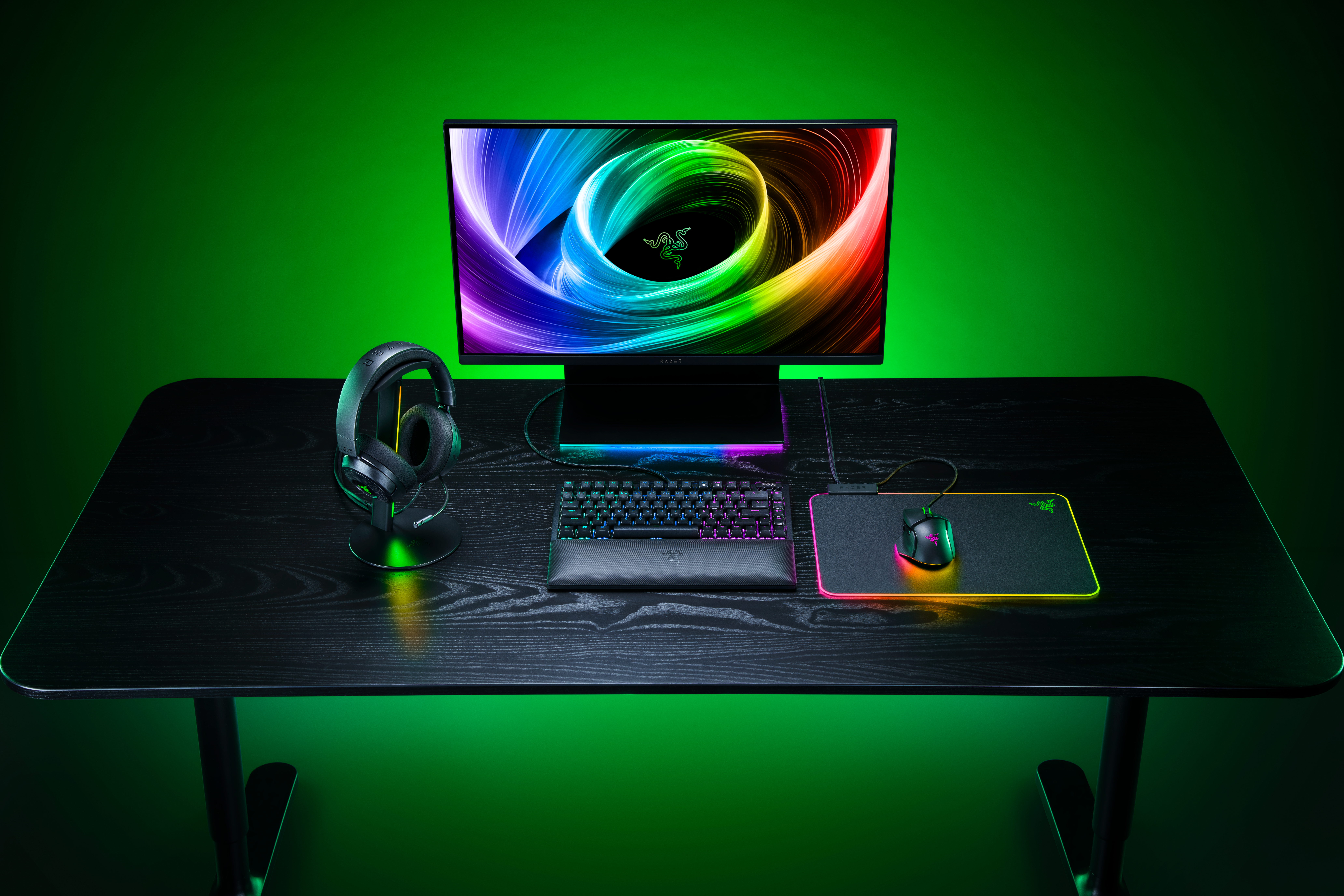 Ein Gaming-Schreibtisch mit Monitor, beleuchteter Tastatur, Maus, Mauspad und Kopfhörern vor grünem Hintergrund.
