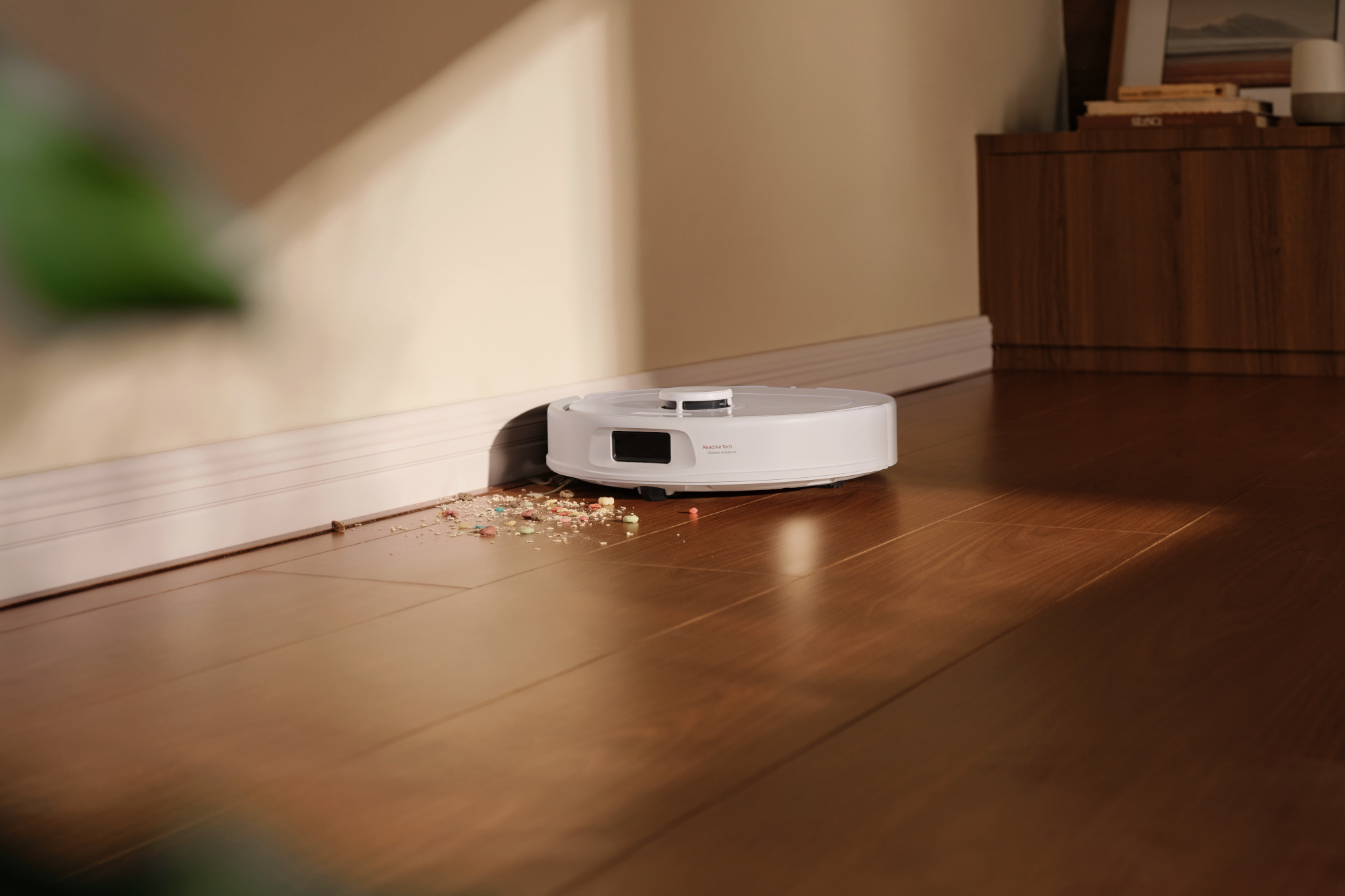 Egy fehér robotporszívó takarítja a fapadlót a szegélylécnél lévő morzsák mellett.