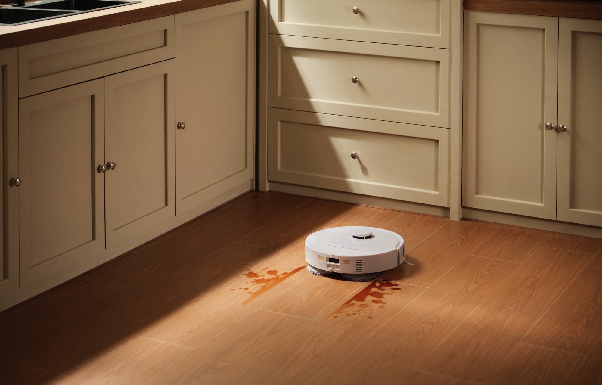 Egy fehér robotporszívó kávéfoltot takarít egy fa padlójú konyhában.